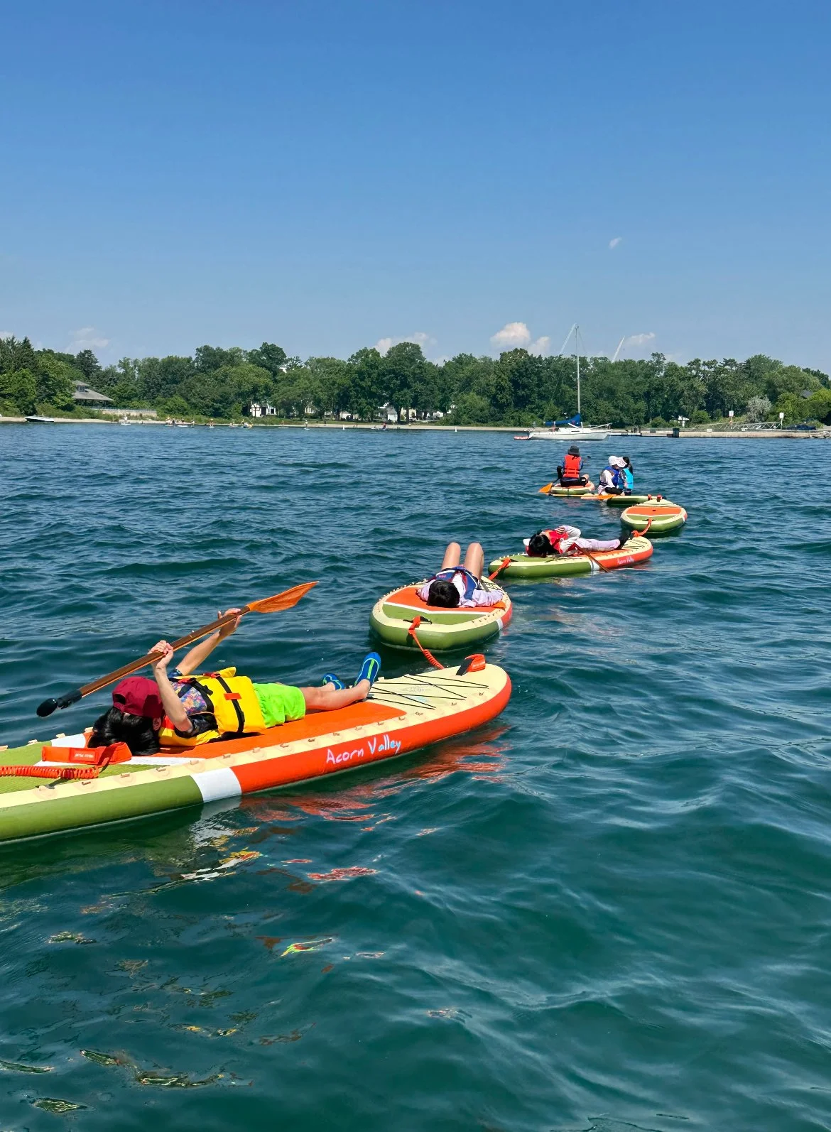 Children in life jackets lying on paddleboards and floating on water, with trees and sailboats in the background on a sunny day.