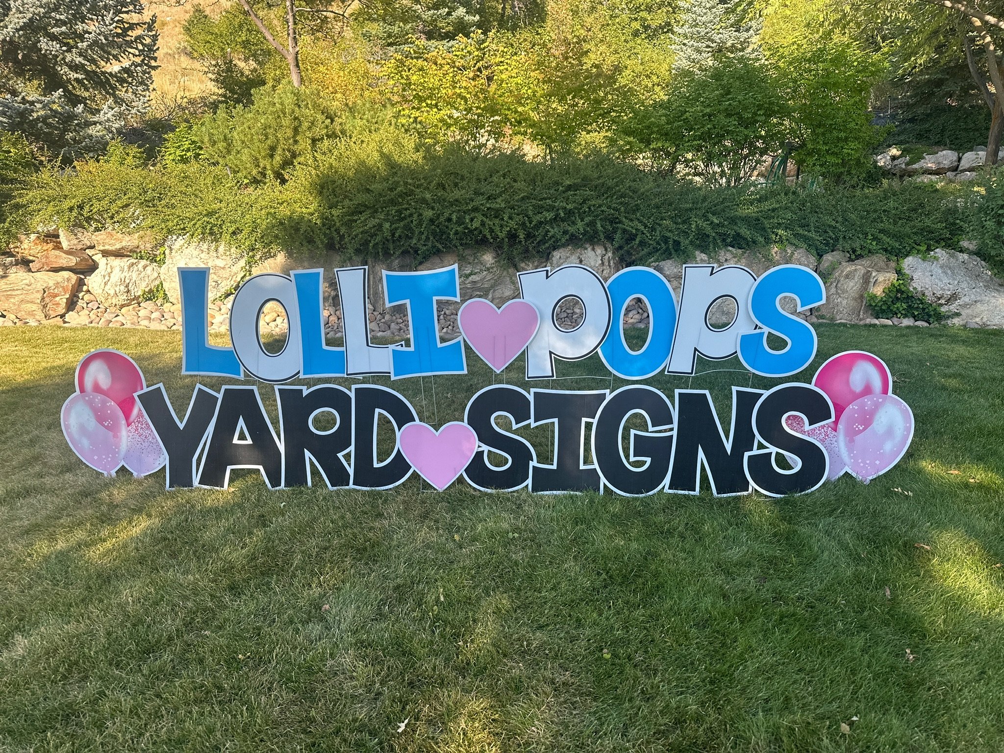 Colorful outdoor sign reading 'Lollipops Yardsigns' with pink balloons and hearts, on a grassy lawn with bushes and rocks in the background.