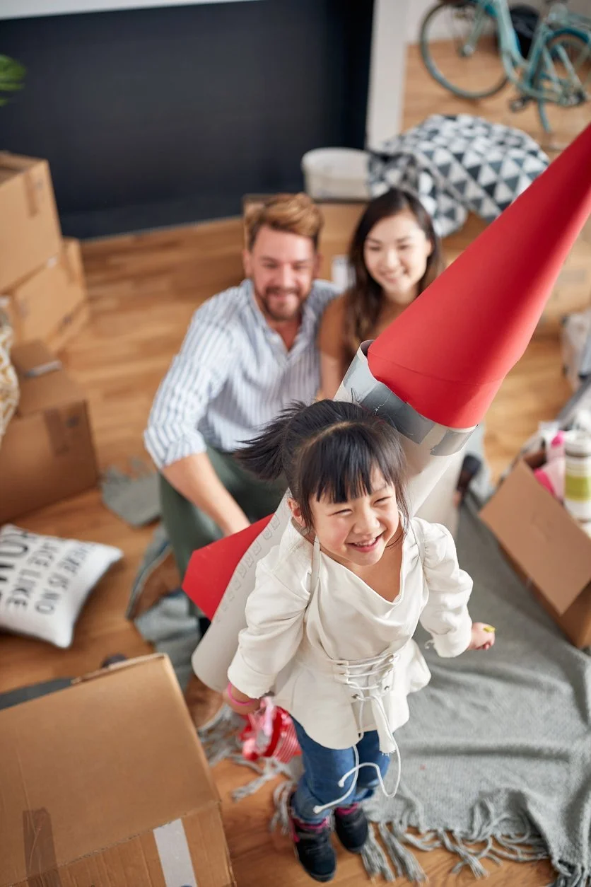 A young girl happy and smiling, wearing a white coat, holding a rolled-up paper rocket, with a man and woman smiling behind her in a cluttered room with cardboard boxes, a pillow, and a bicycle in the background.