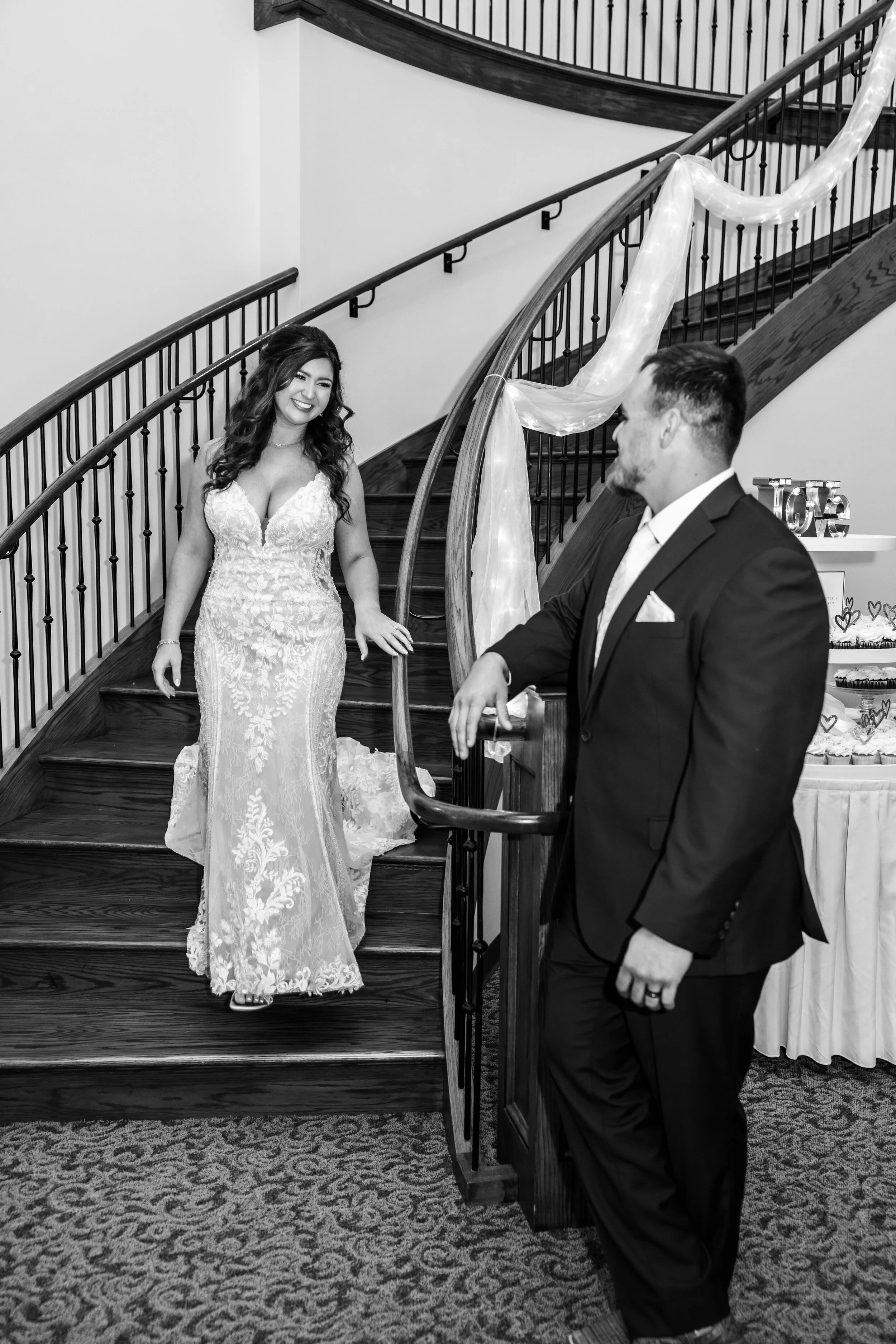 A woman in a wedding dress and a man in a suit smiling at each other on a staircase decorated with sheer fabric for a wedding celebration.
