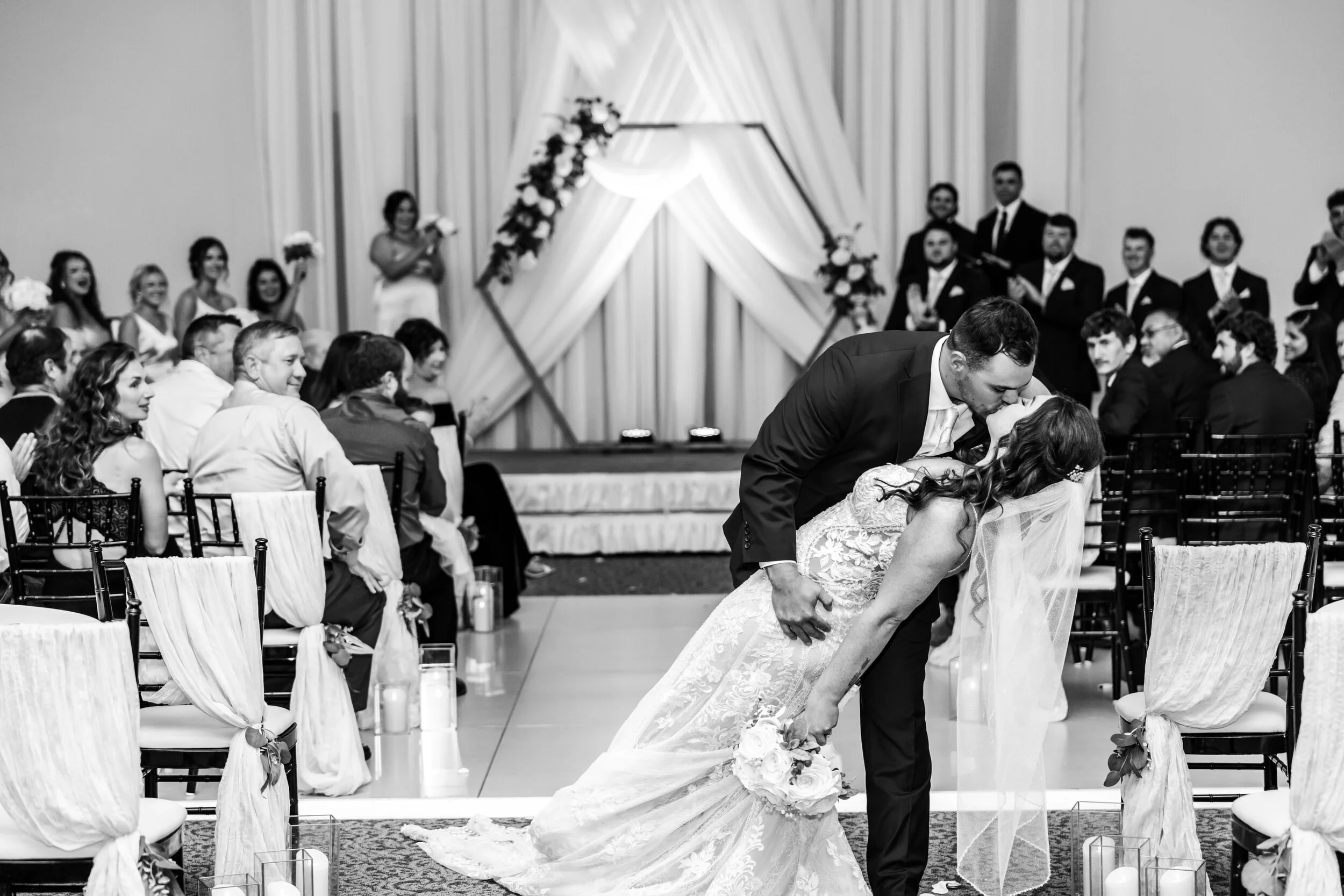 A couple sharing a kiss during their wedding ceremony with guests and bridesmaids in the background.