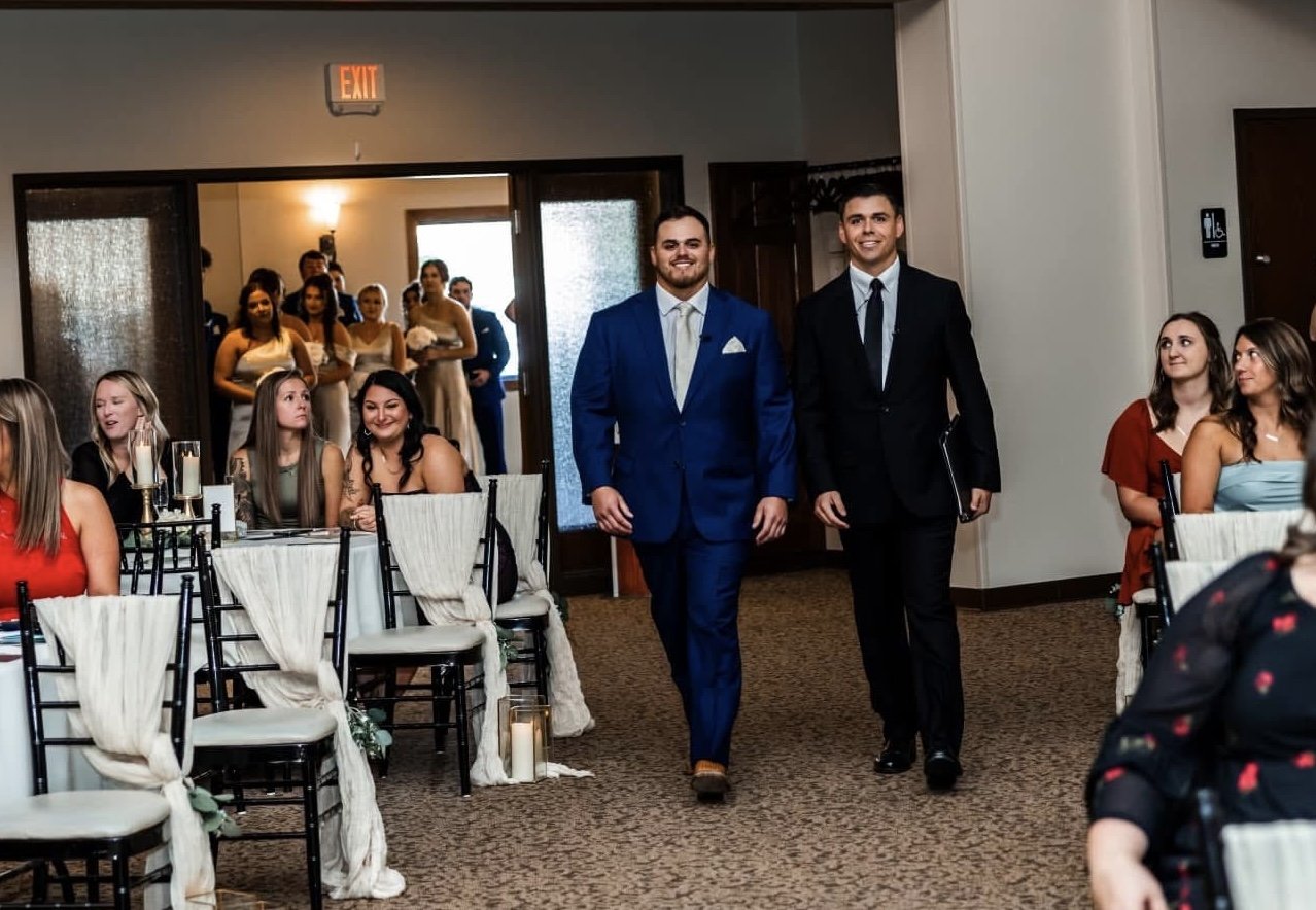 Two men in suits walking into a wedding reception, with guests seated at tables. The room has a warm ambiance with candles and draped chairs, and a group of bridesmaids and guests in the background.