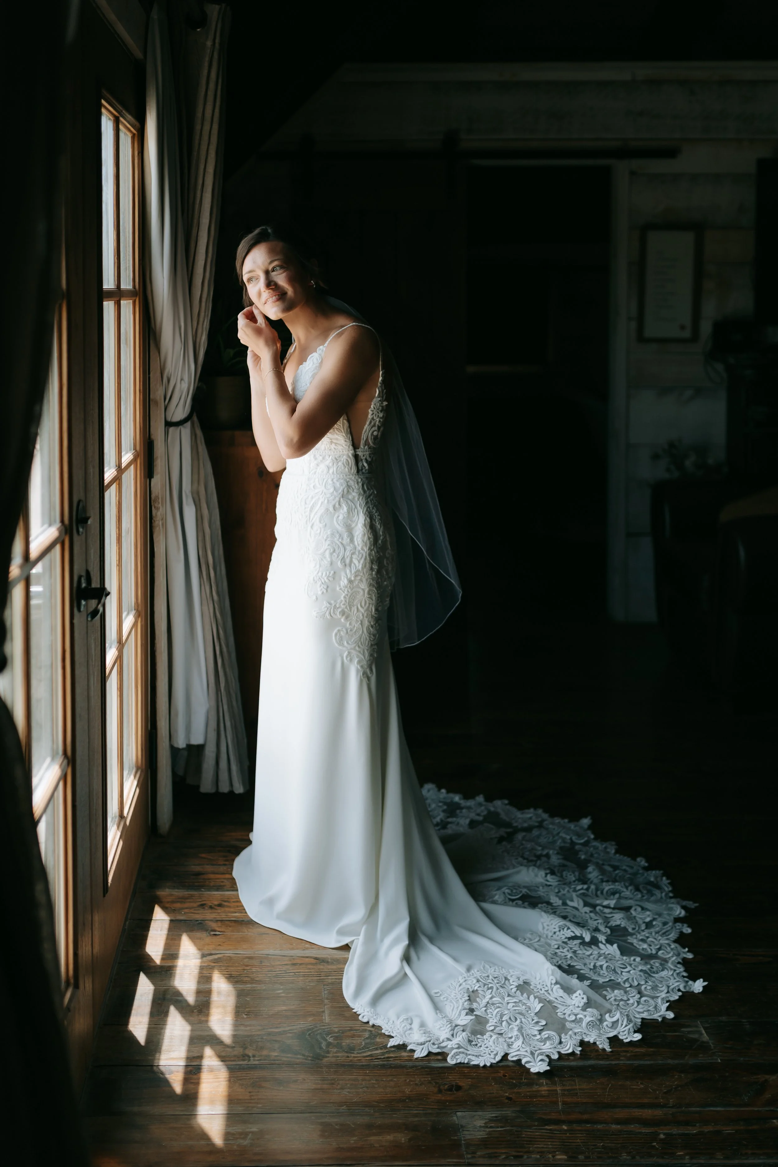 A bride in a white lace wedding gown looks out the window, smiling softly, with sunlight casting shadows on the wooden floor.