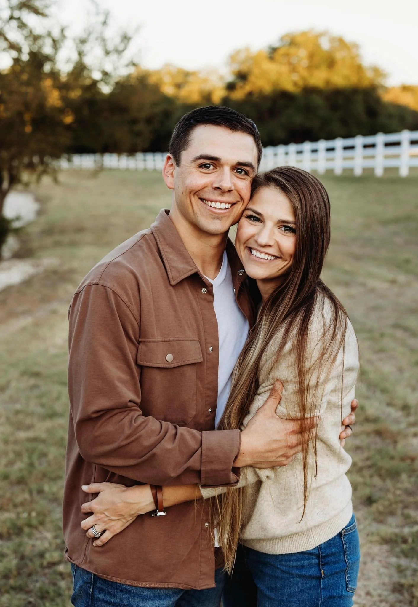 A smiling couple embraces outdoors in a grassy area with trees and a white fence in the background during sunset.