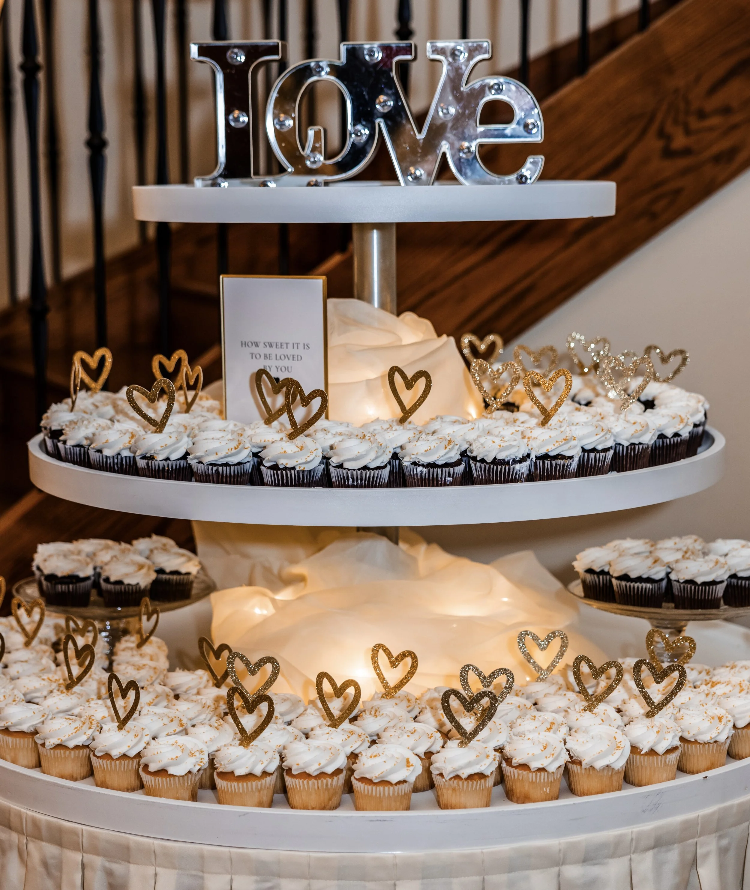 A three-tier dessert display with cupcakes on the bottom and middle tiers, topped with golden heart decorations and a silver "LOVE" sign on the top tier. A small sign with a quote is placed on the middle tier.