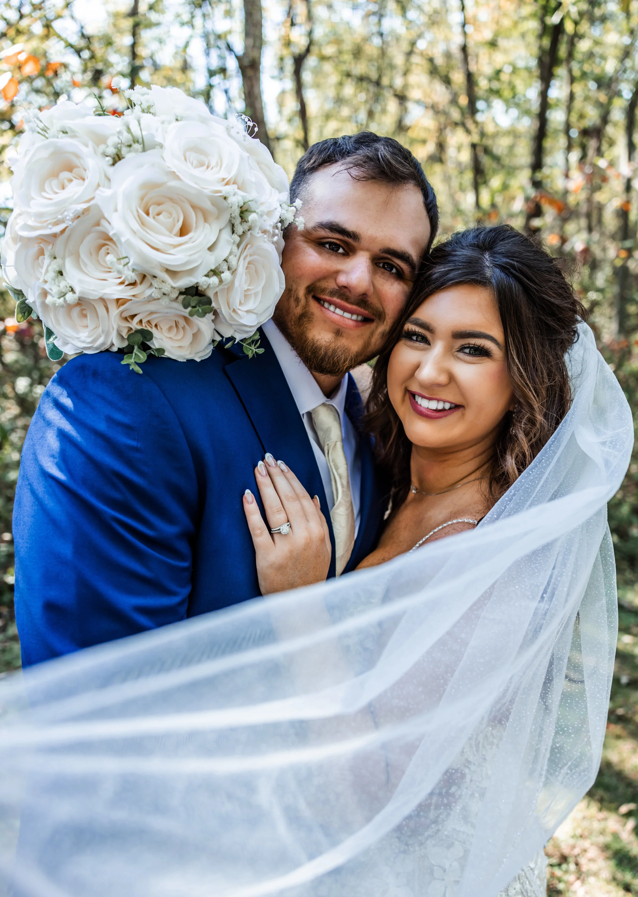A newlywed couple smiling, embracing outdoors with trees in the background, the bride holding a bouquet of white roses and wearing a veil, and the groom in a blue suit.