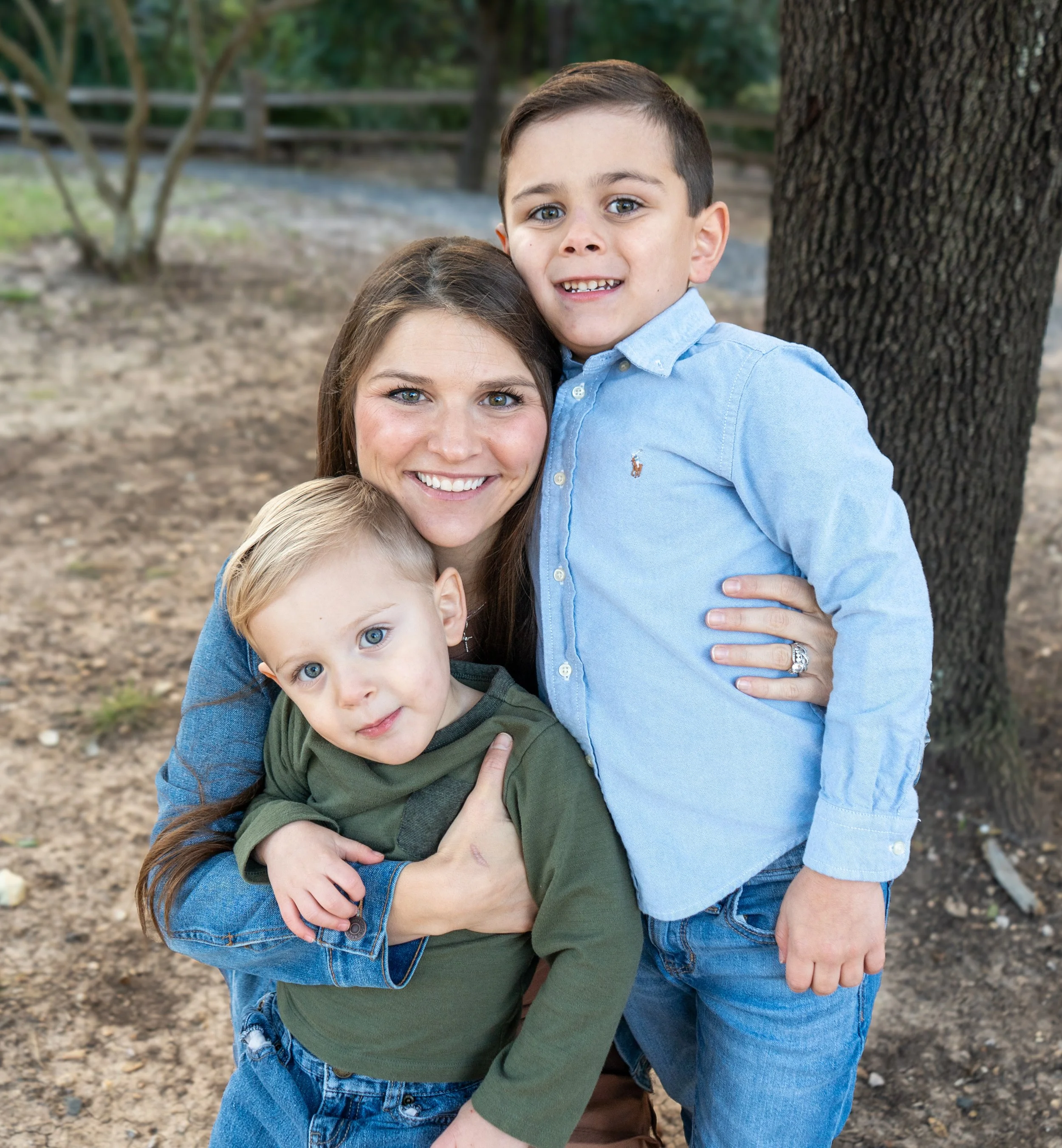 A woman with long brown hair smiling, holding two young boys outdoors near a tree and fence. The boy on the right has dark hair and blue eyes, wearing a light blue shirt. The boy on the left has blond hair and blue eyes, wearing an olive green shirt. All are smiling and close together.