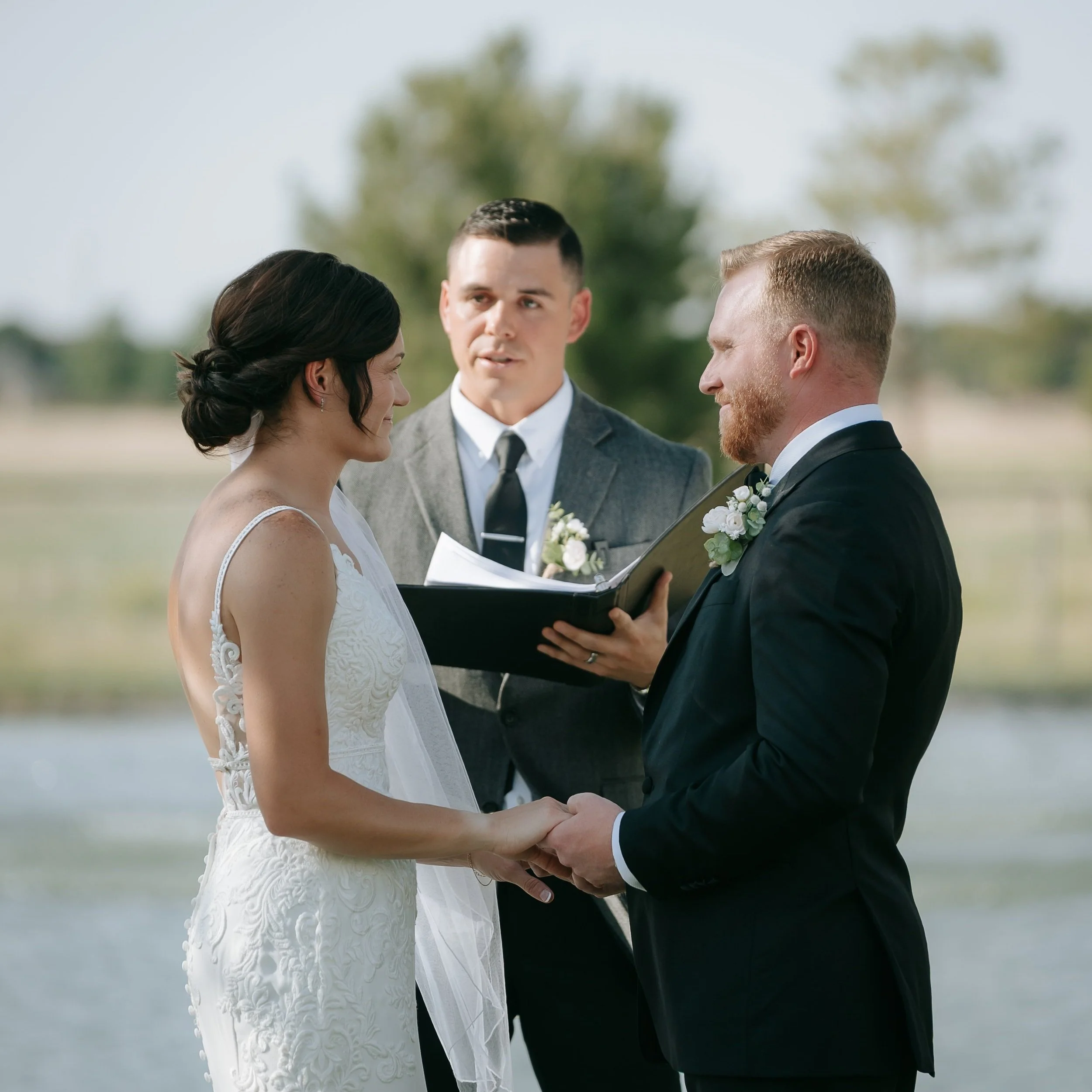 A couple getting married and holding hands during their wedding ceremony outdoors with an officiant standing behind them, holding a folder and reading vows, in a scenic outdoor setting with trees and water in the background.