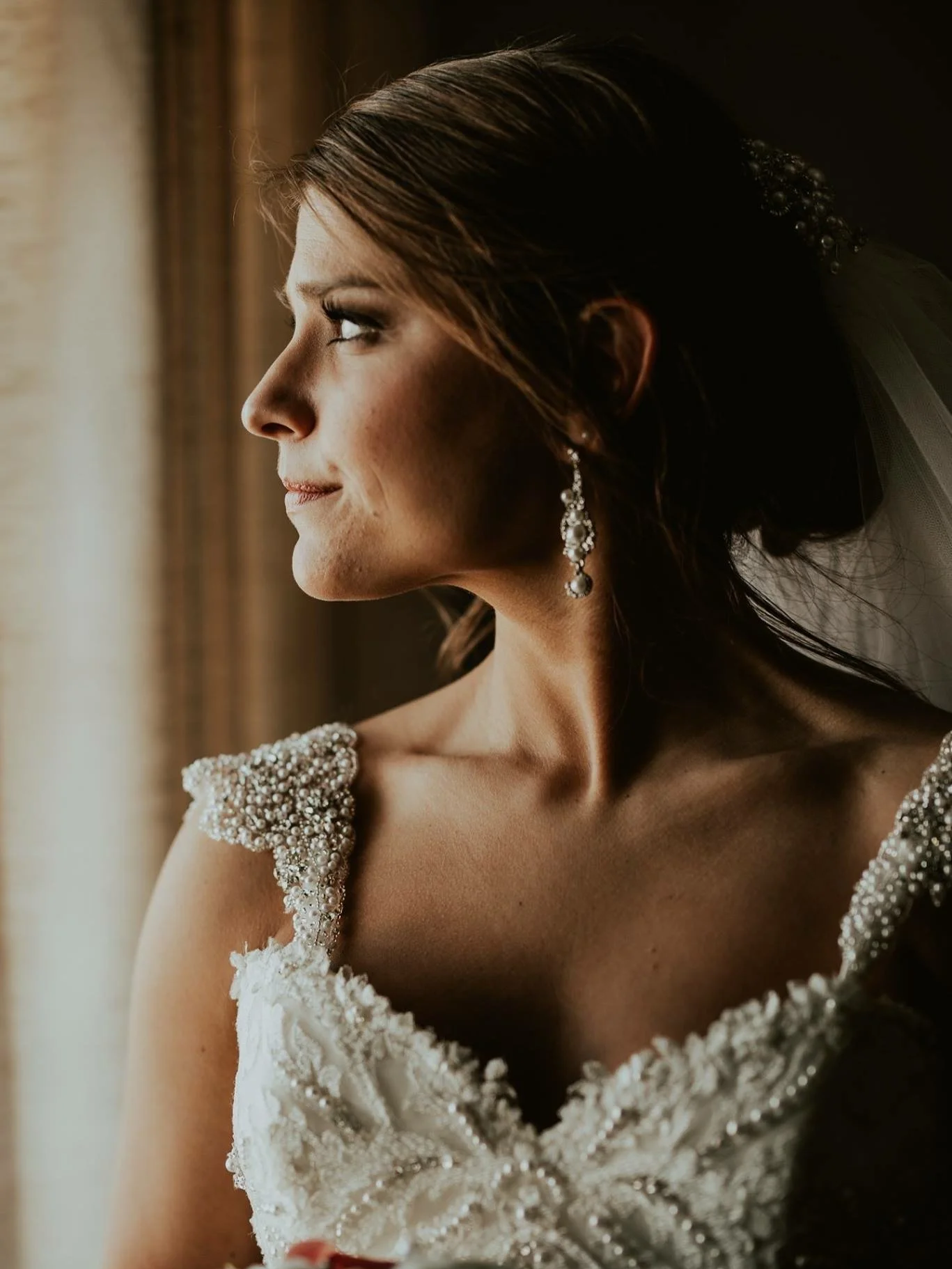A bride looking out a window, wearing a wedding dress with pearl embellishments and dangling earrings, with her hair styled in an elegant updo.