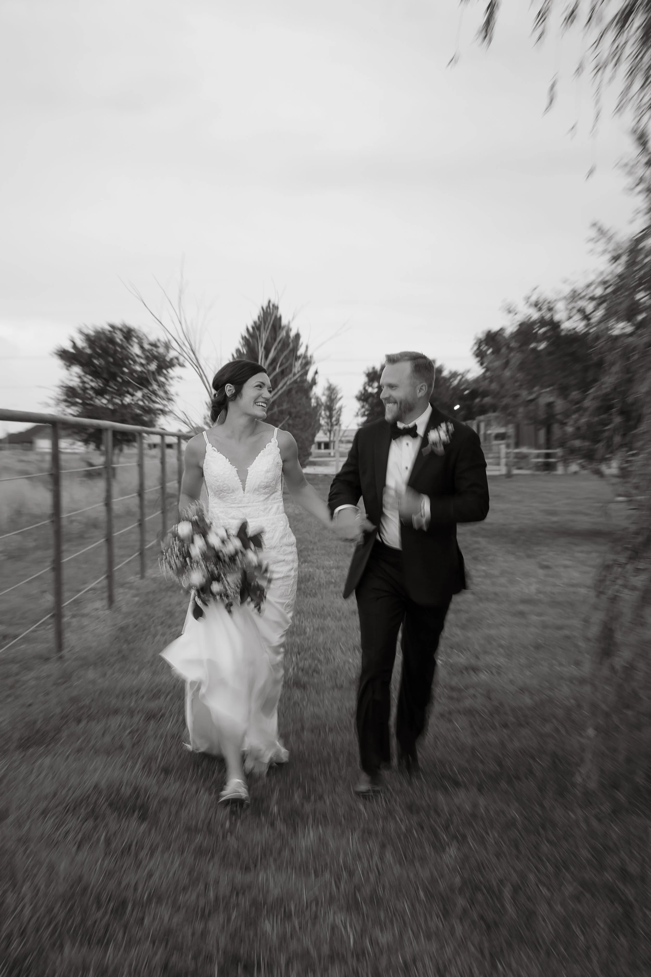 Black and white photo of a bride and groom running outside, holding hands and smiling, with trees and fencing in the background.