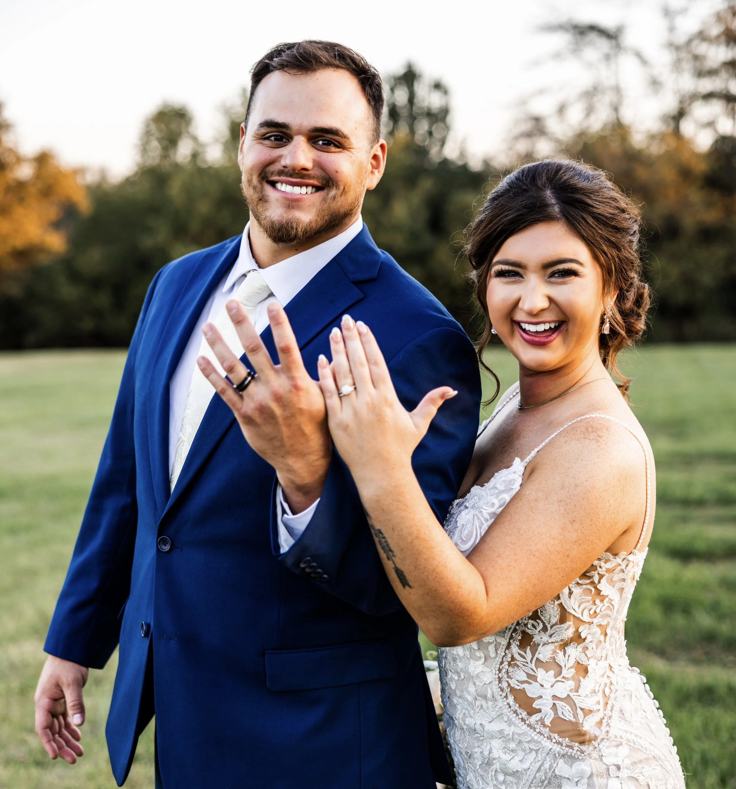 A smiling man and woman in wedding attire displaying their rings outdoors in a grassy area with trees in the background.