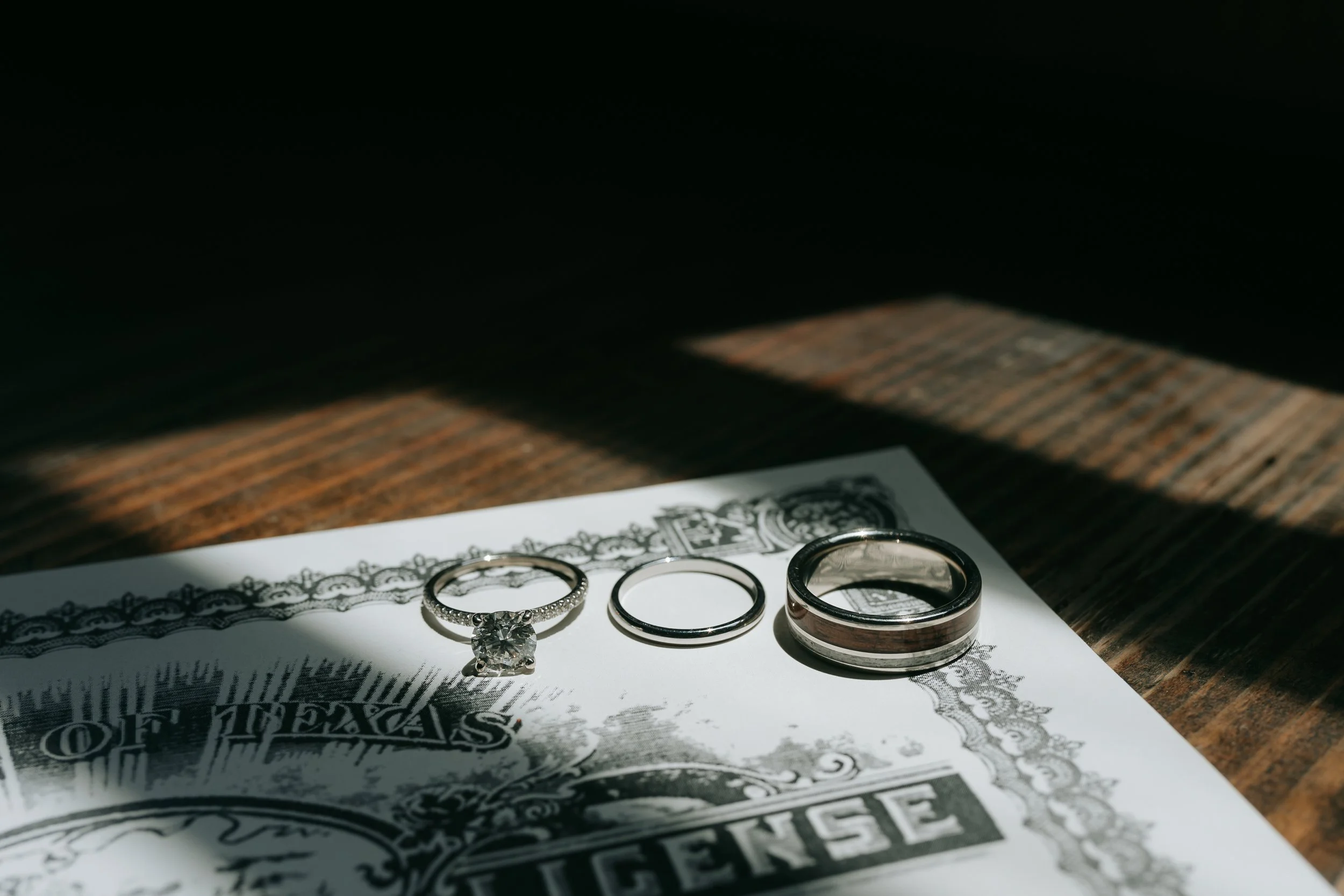 Diamond engagement ring, wedding band, and men's wedding band placed on a paper with a decorative border and text, illuminated by natural light on a wooden surface.