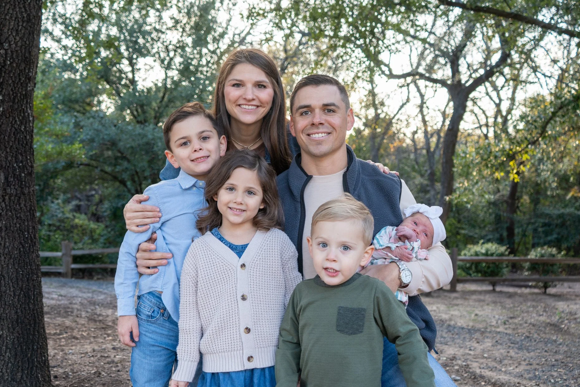 A happy family of six, including two adults and five children, standing outdoors in a park with trees and sunlight behind them.