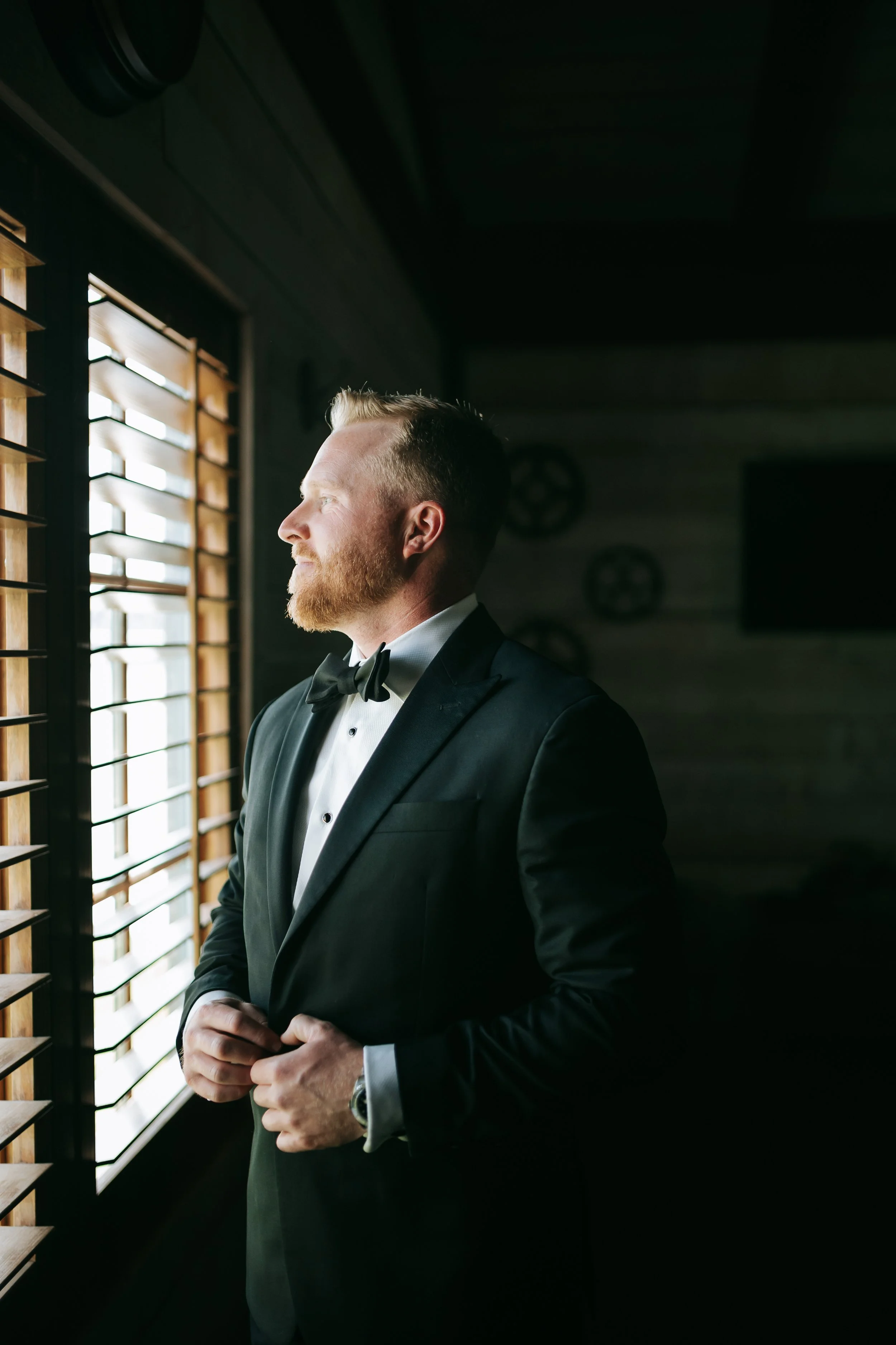 A groom in a black tuxedo and bow tie standing inside, looking out a window with wooden blinds.