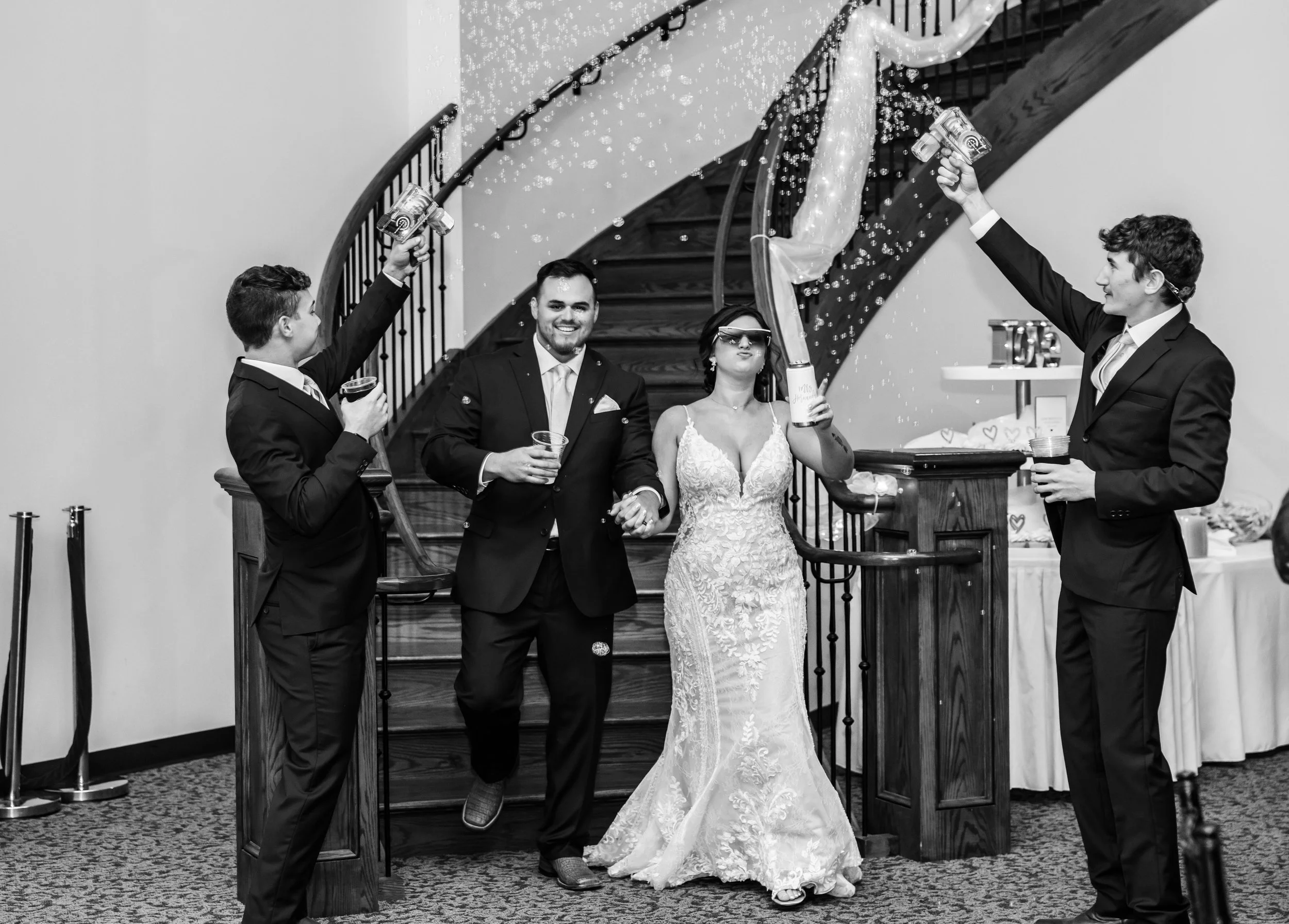 A black and white photo of a wedding celebration with five people. A bride in a lace wedding gown is in the center, wearing protective glasses and holding a large confetti popper. Flanking her are two men in suits, one holding a glass, and two men on either side, each wearing suits and holding small bottles, spraying confetti from the bottles. They are gathered near a staircase in an indoor setting.