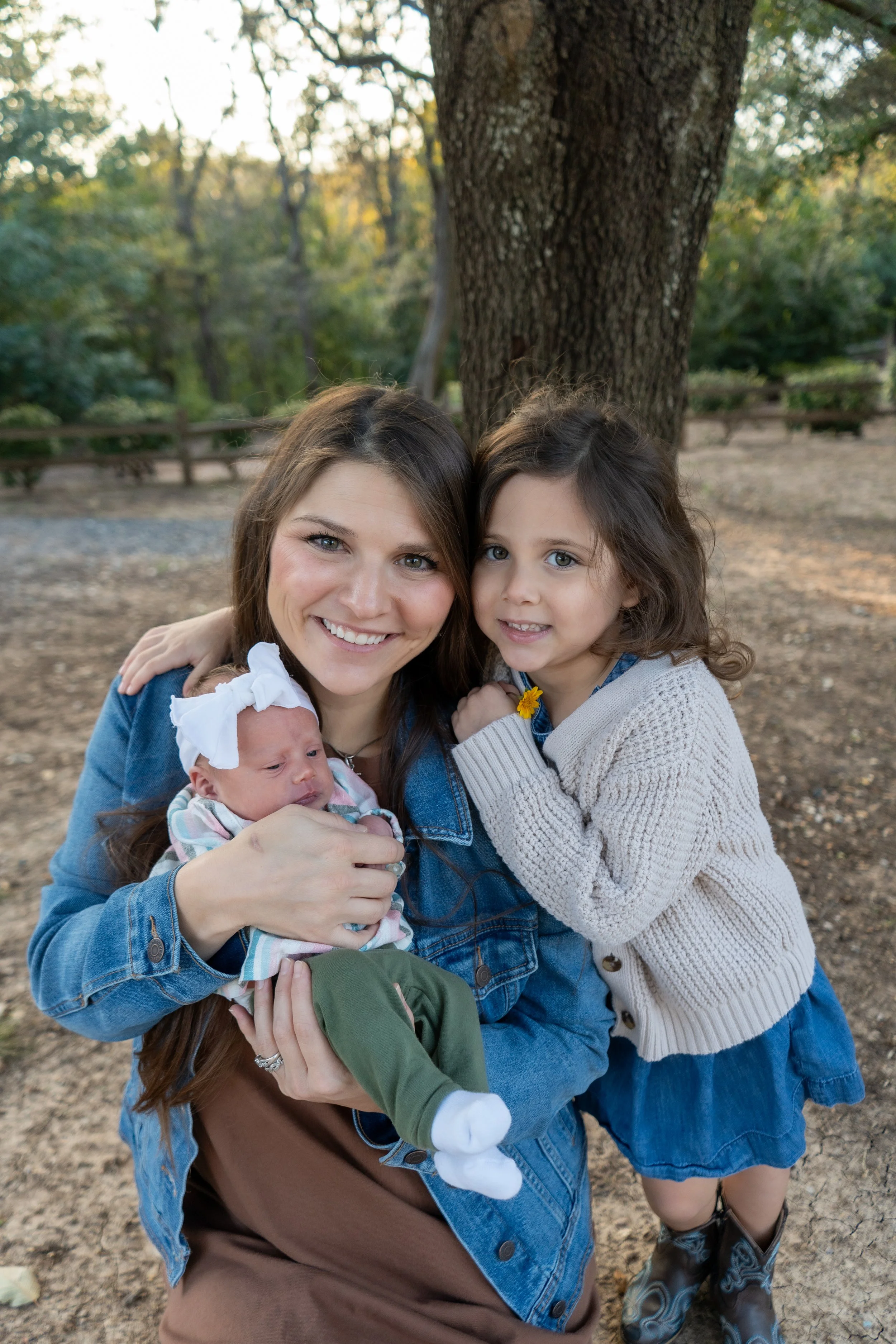 A woman with two young girls, one older and one a newborn, posing outdoors near a large tree in a park.