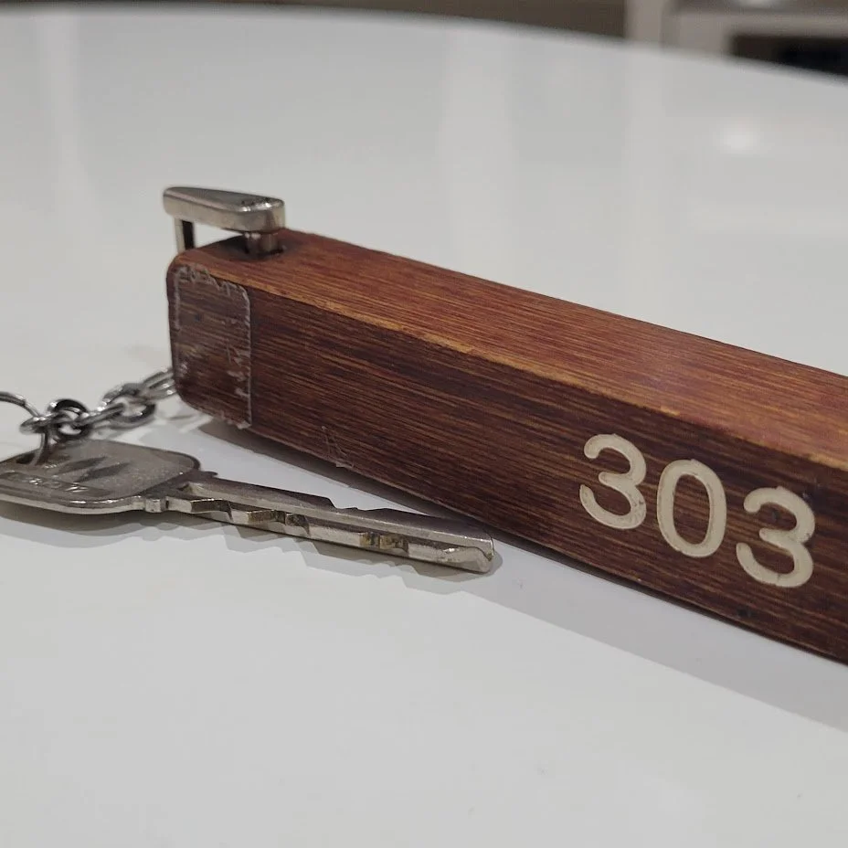 A wooden keychain with the number 303 engraved on it, attached to a metal key with a chain. The keychain has a metal clip on one end.