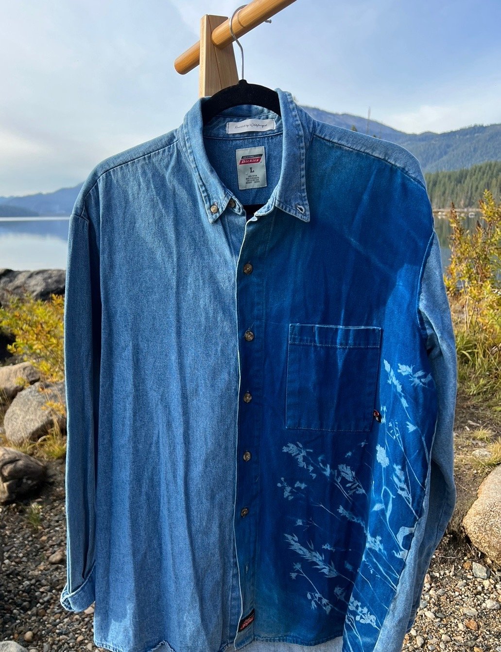 A denim shirt hanging on a wooden hanger outside near a body of water with mountains in the background.