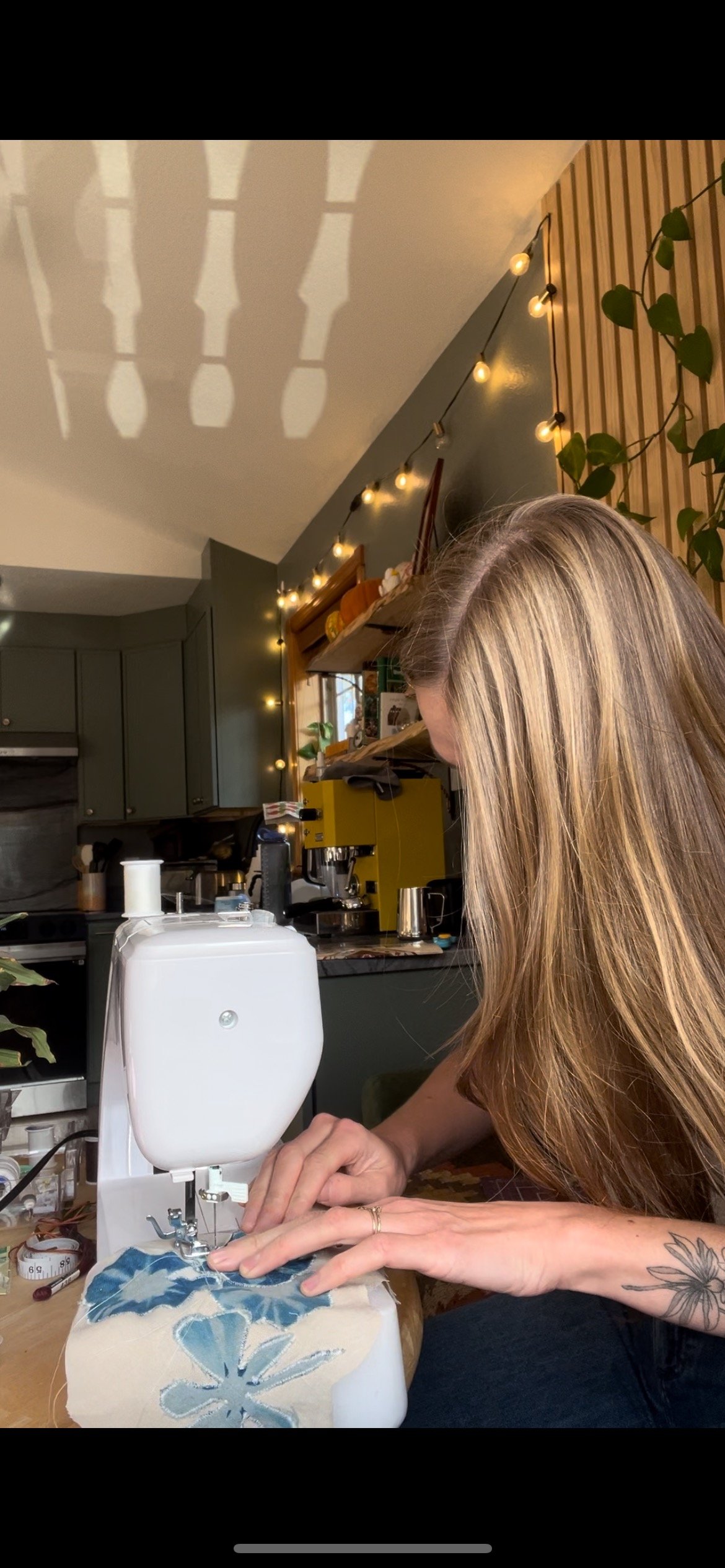 A person sewing fabric on a white sewing machine in a cozy kitchen decorated with string lights and green plants.