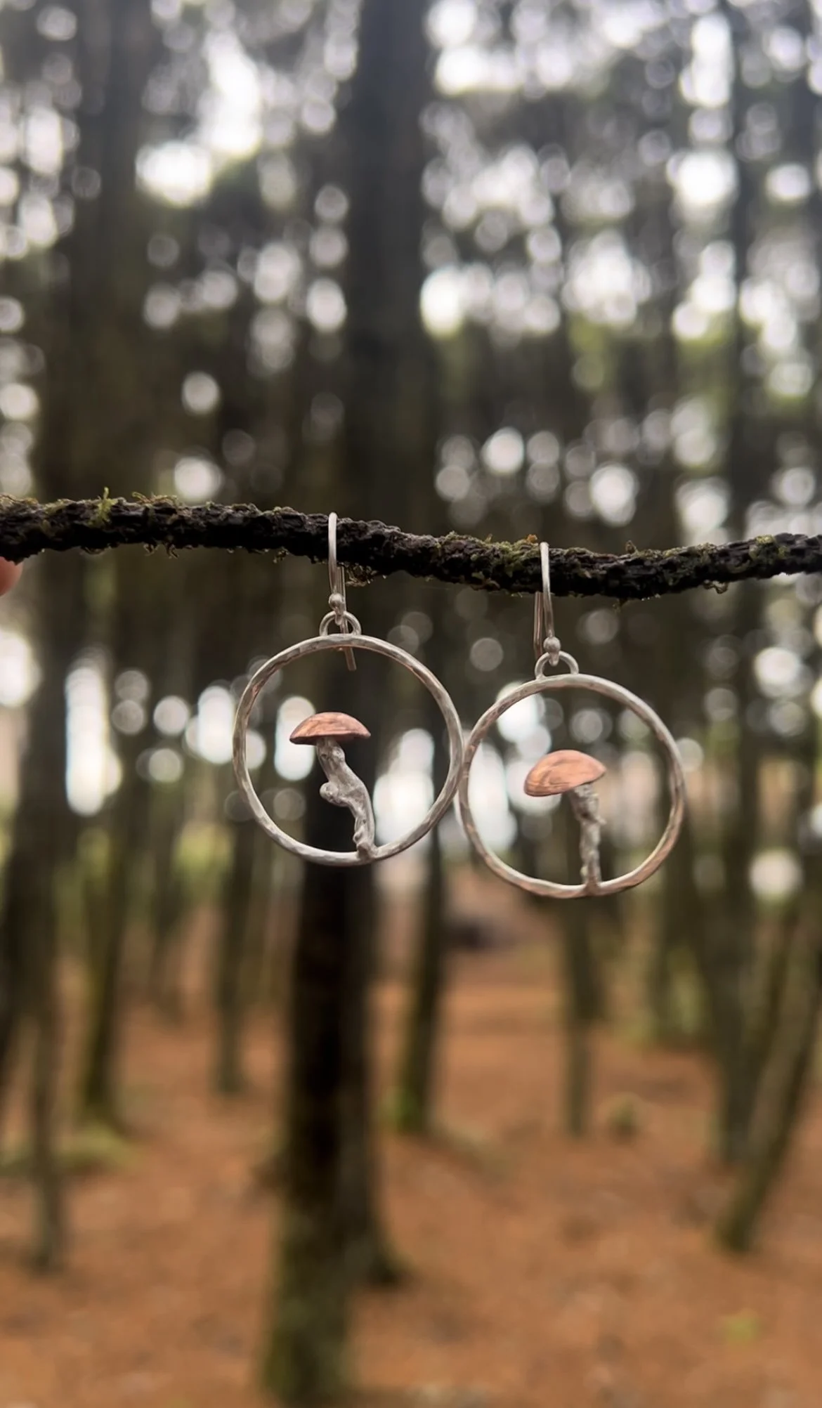 Pair of earrings hanging on a tree branch in a forest with blurred trees and leaves in the background.
