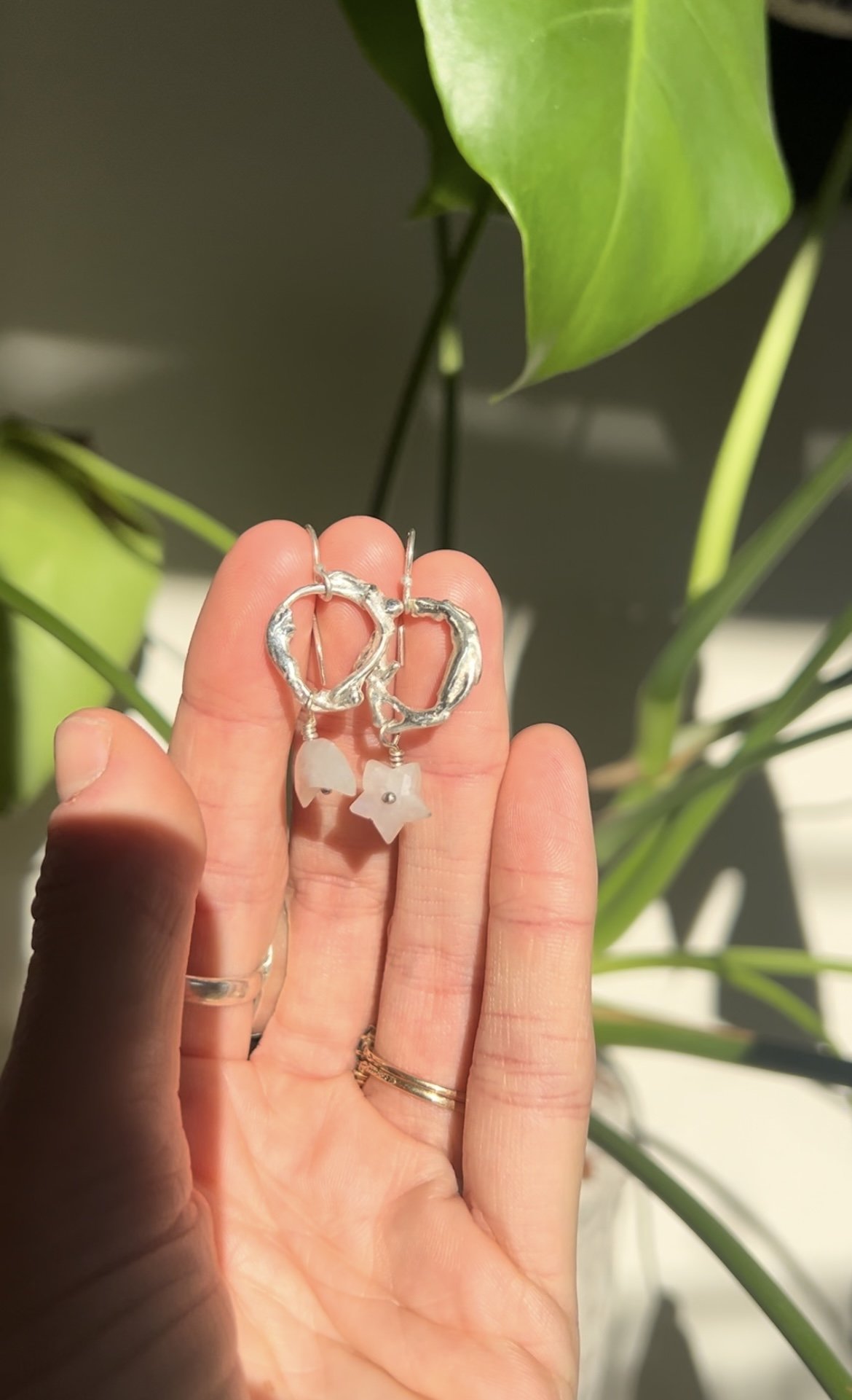Silver hoop earrings with small charms, held in a person's hand, with green leafy plants in the background.