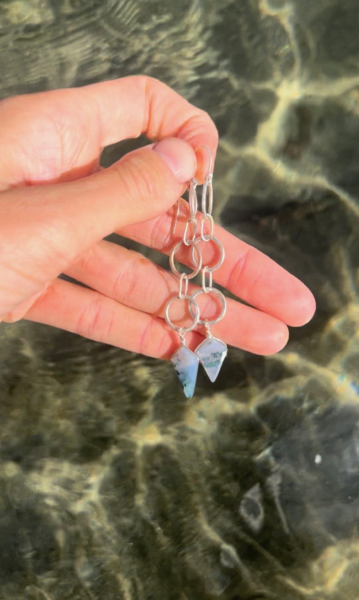 Hand holding three pairs of silver earrings with arrowhead-shaped stones against a rocky background.