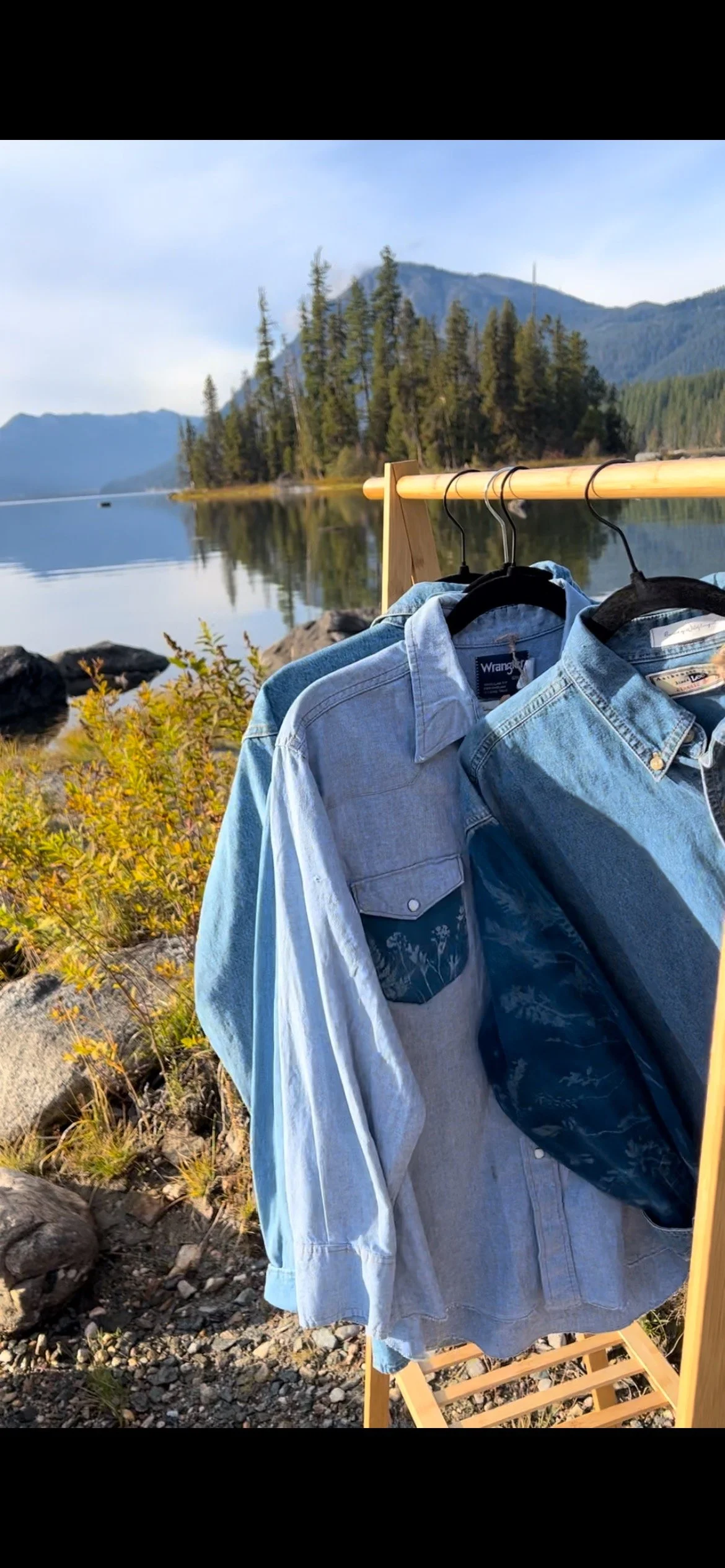 Three denim shirts hanging on a clothing rack near a lakeside with mountains and trees in the background.