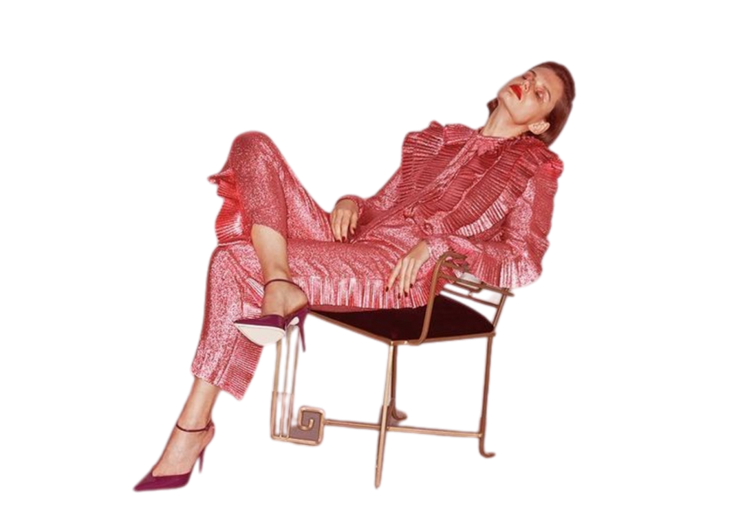 A woman wearing a shiny red patterned suit with a ruffled top sitting on a modern black and gold chair, with one leg crossed over the other, looking to the side with a confident expression.