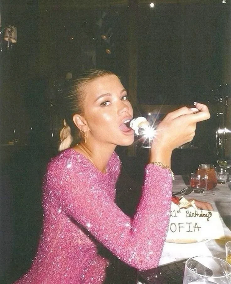 A woman with blonde hair styled in a low ponytail wearing a sparkly pink dress, sitting at a table, eating cake, with a birthday cake in front of her that says 'Happy 1st Birthday Sofia.'