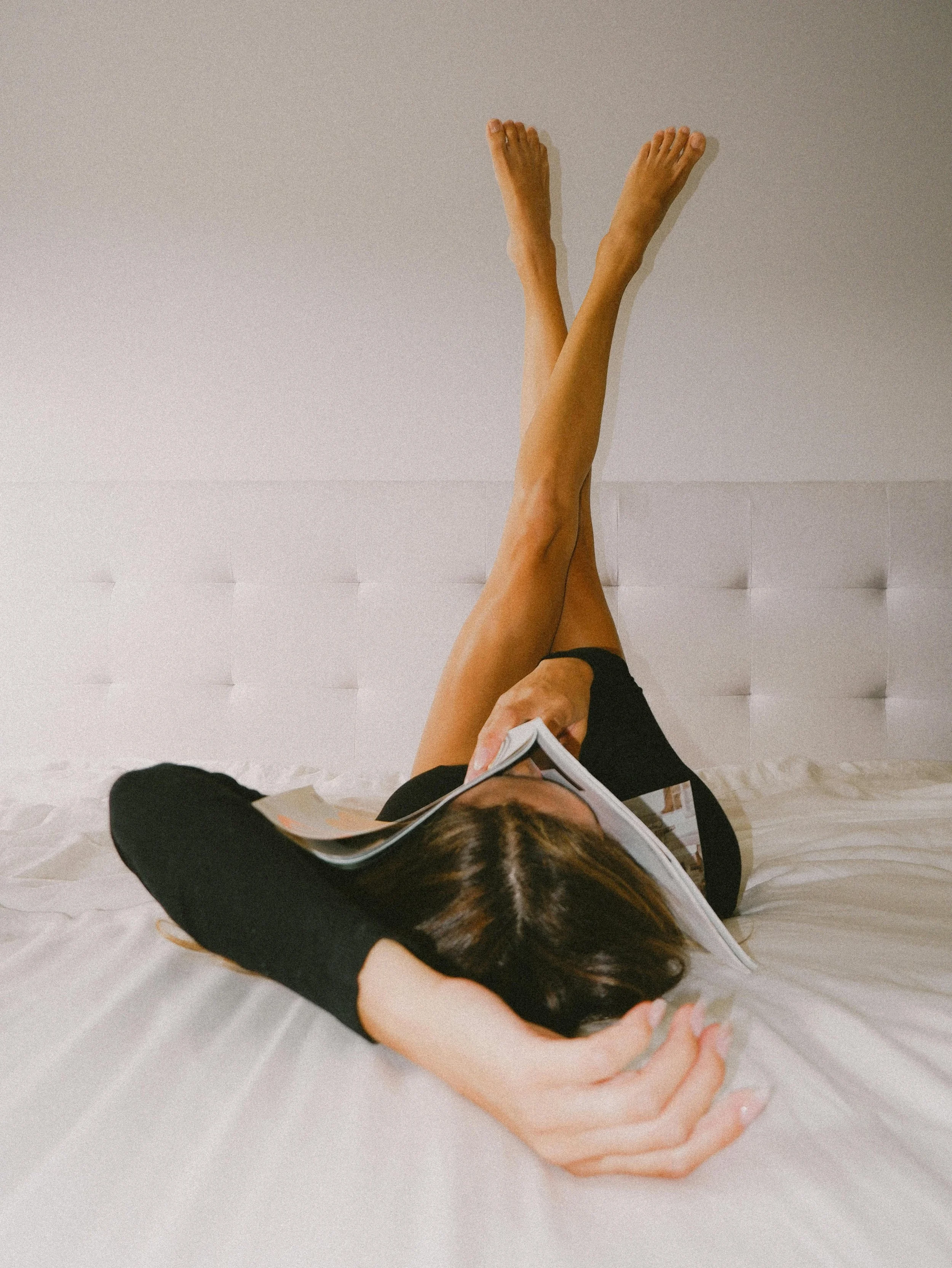 A woman lying on a bed, reading a book with her legs raised and resting against the wall.