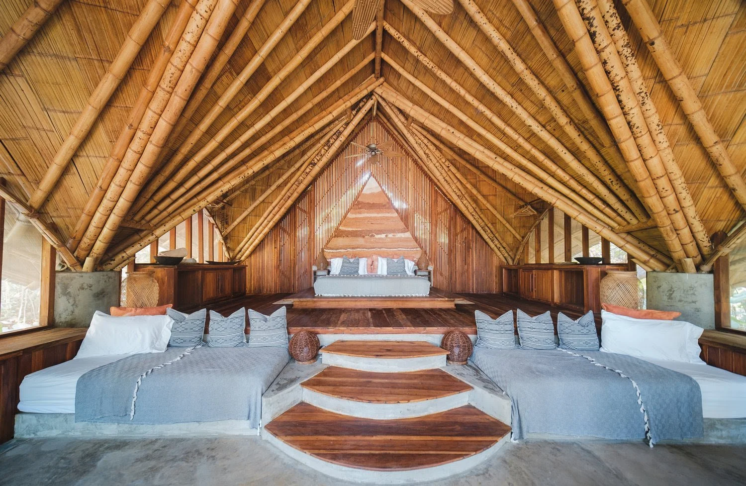 Interior of a rustic, wooden A-frame bedroom with two beds on the lower level and a raised bed on the upper level, all with gray bedding and decorative pillows, surrounded by bamboo and wooden paneling.