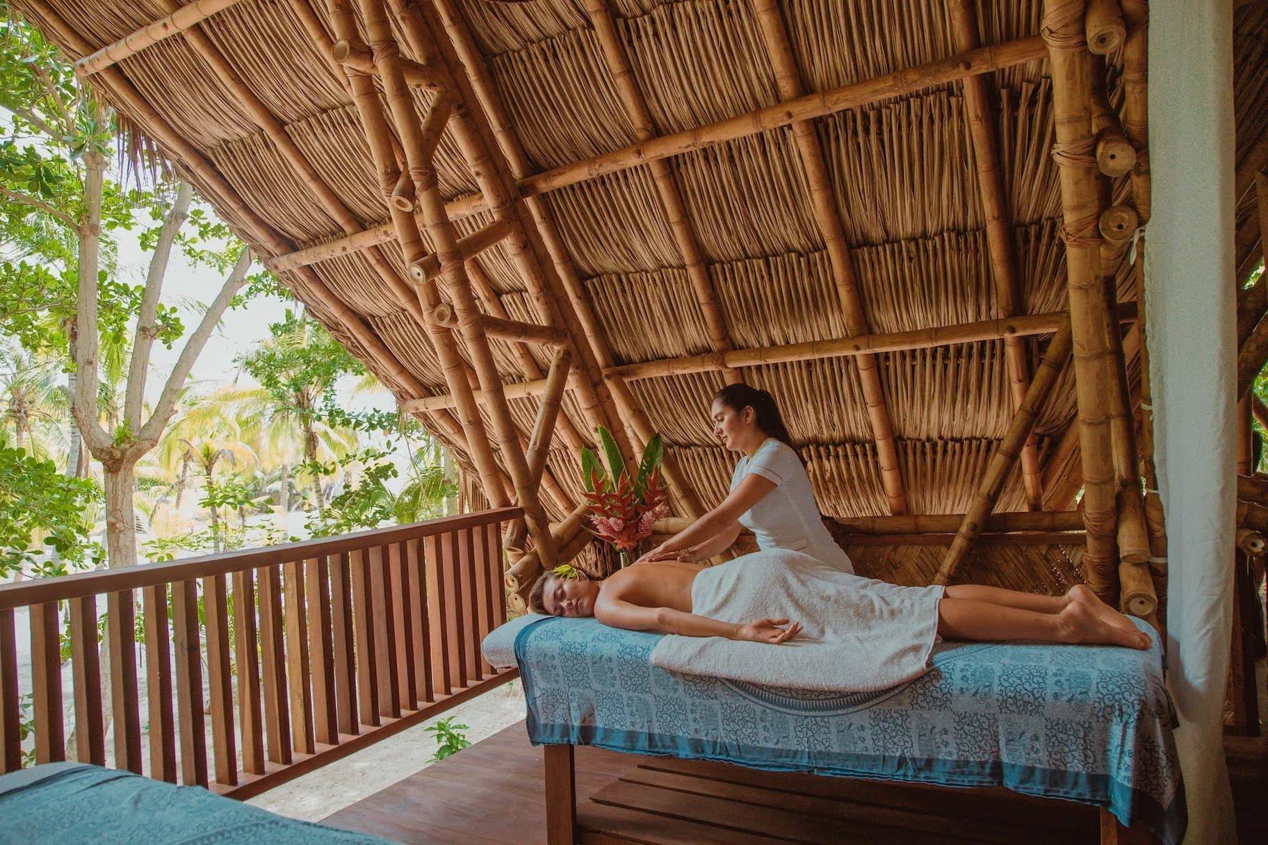 A woman receiving a massage outdoors in a bamboo hut with lush green trees visible outside.