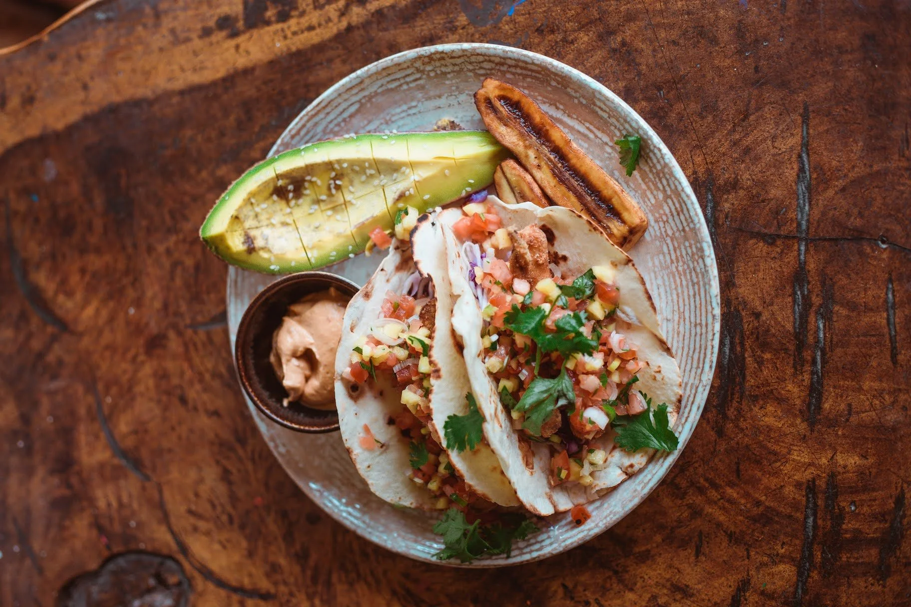 Two fish tacos topped with chopped vegetables and cilantro, served with sliced avocado, grilled plantain, and dipping sauce on a ceramic plate.