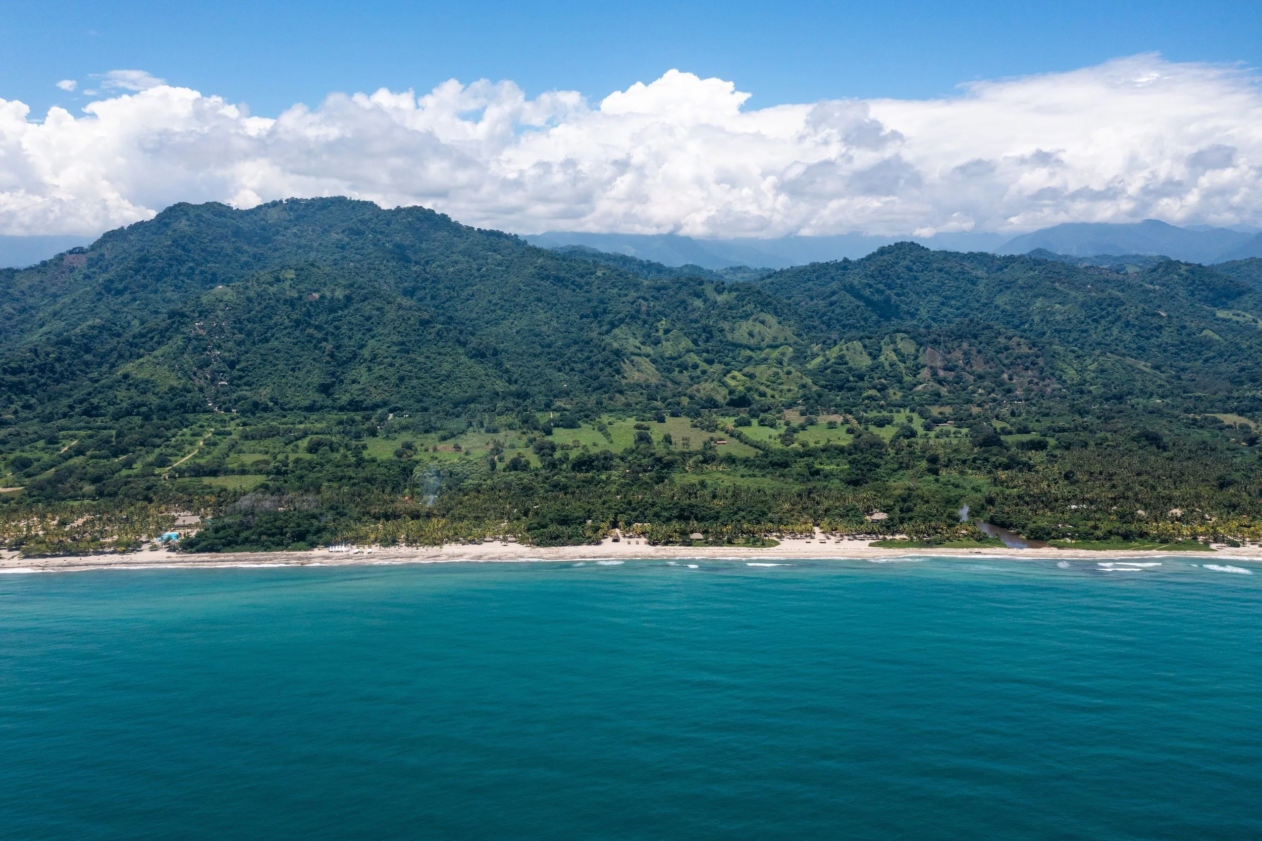 A coastal view showing a green mountain range meeting the ocean with a beach in the foreground and partly cloudy sky.