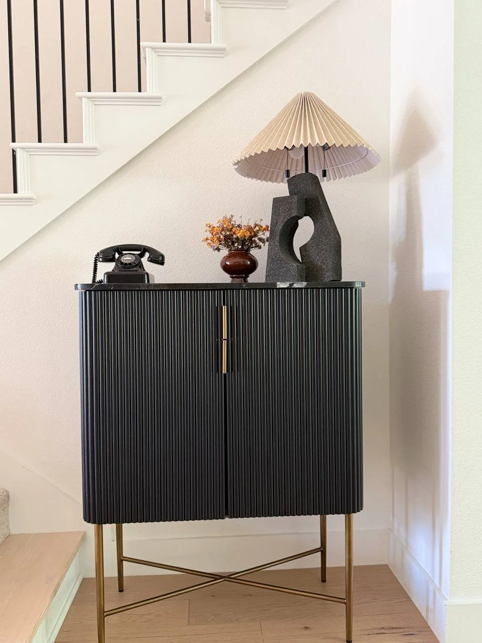 Black fluted cabinet with marble top in an entryway, styled with a vintage rotary telephone, dried flowers, and a sculptural stone lamp