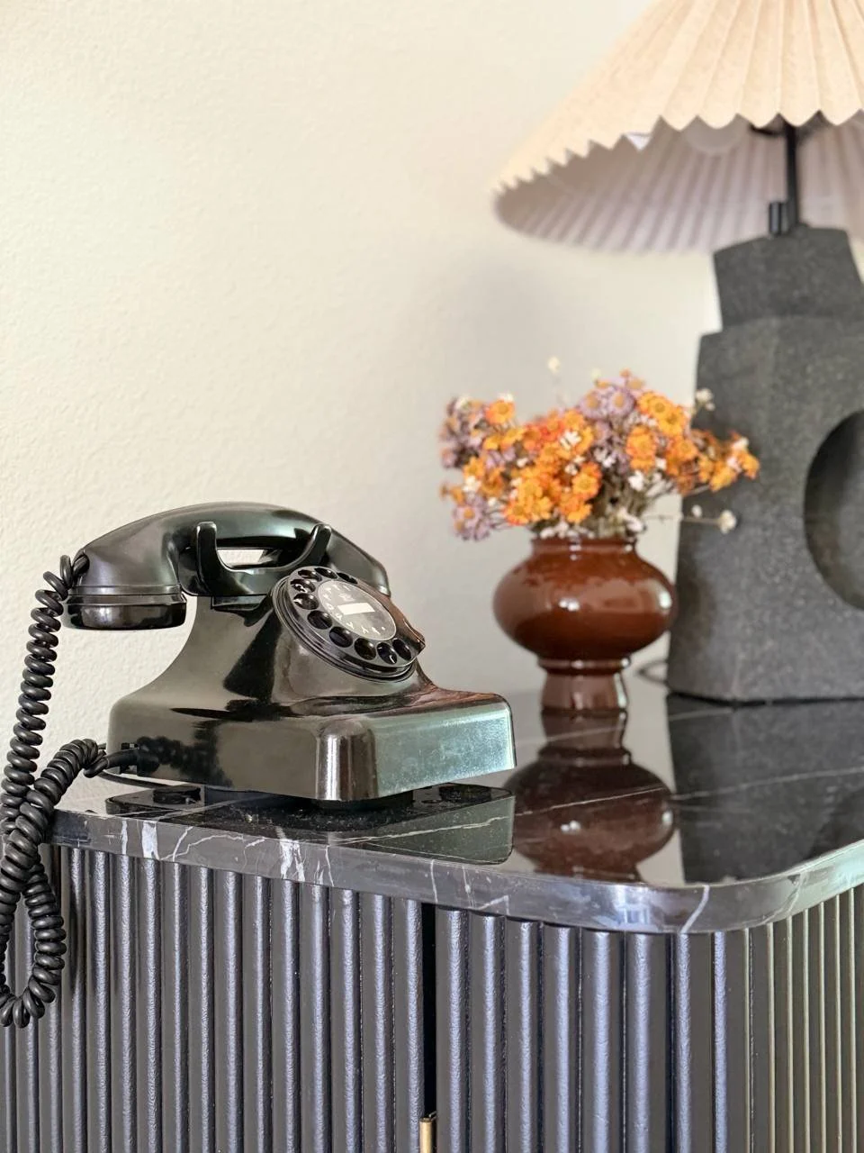 Vintage black rotary telephone on a black marble surface beside dried flowers in a brown ceramic vase