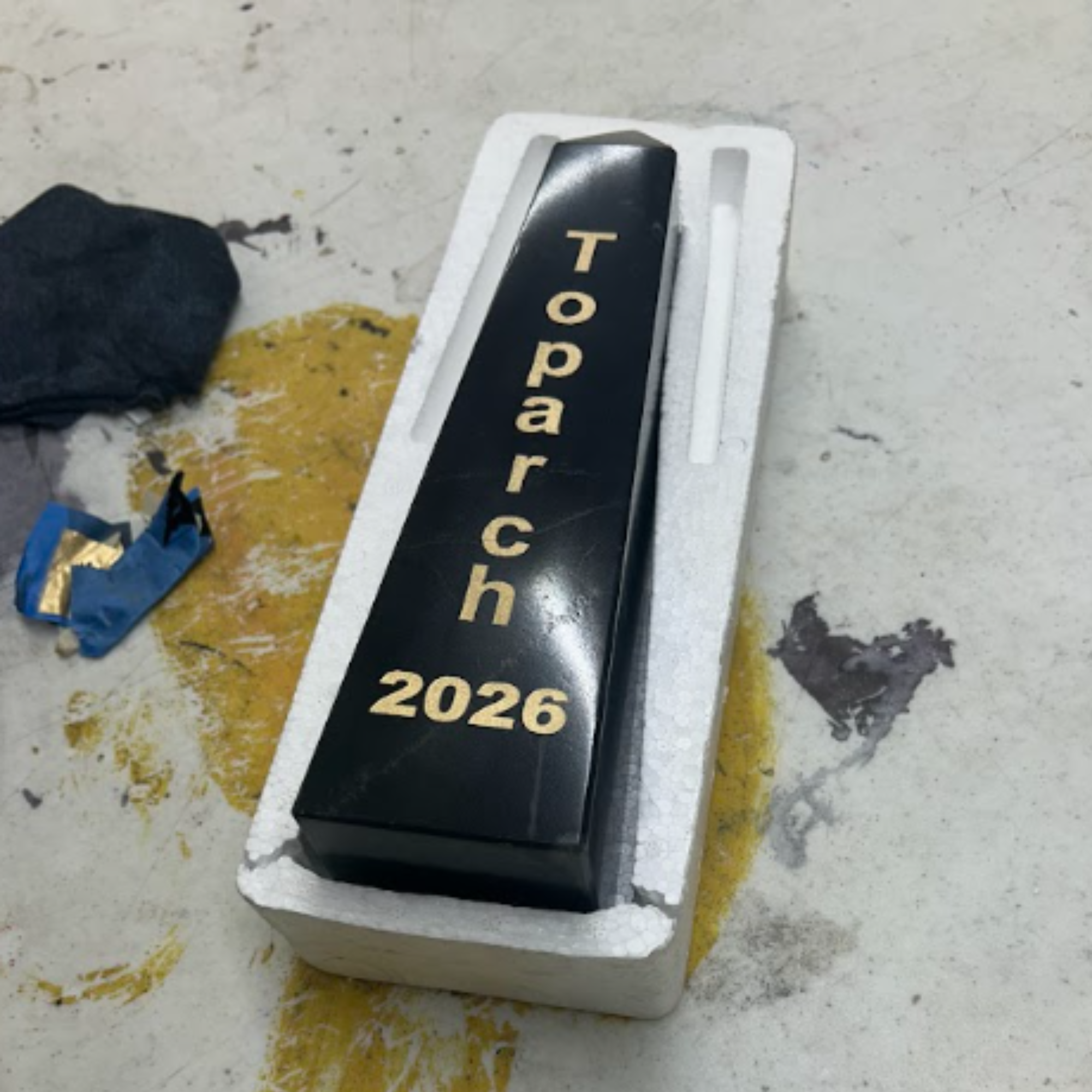 Close-up of a car registration sticker with the year 2026, partially peeled off from a white surface on the ground, next to blackened debris and a small piece of crumpled blue paper.