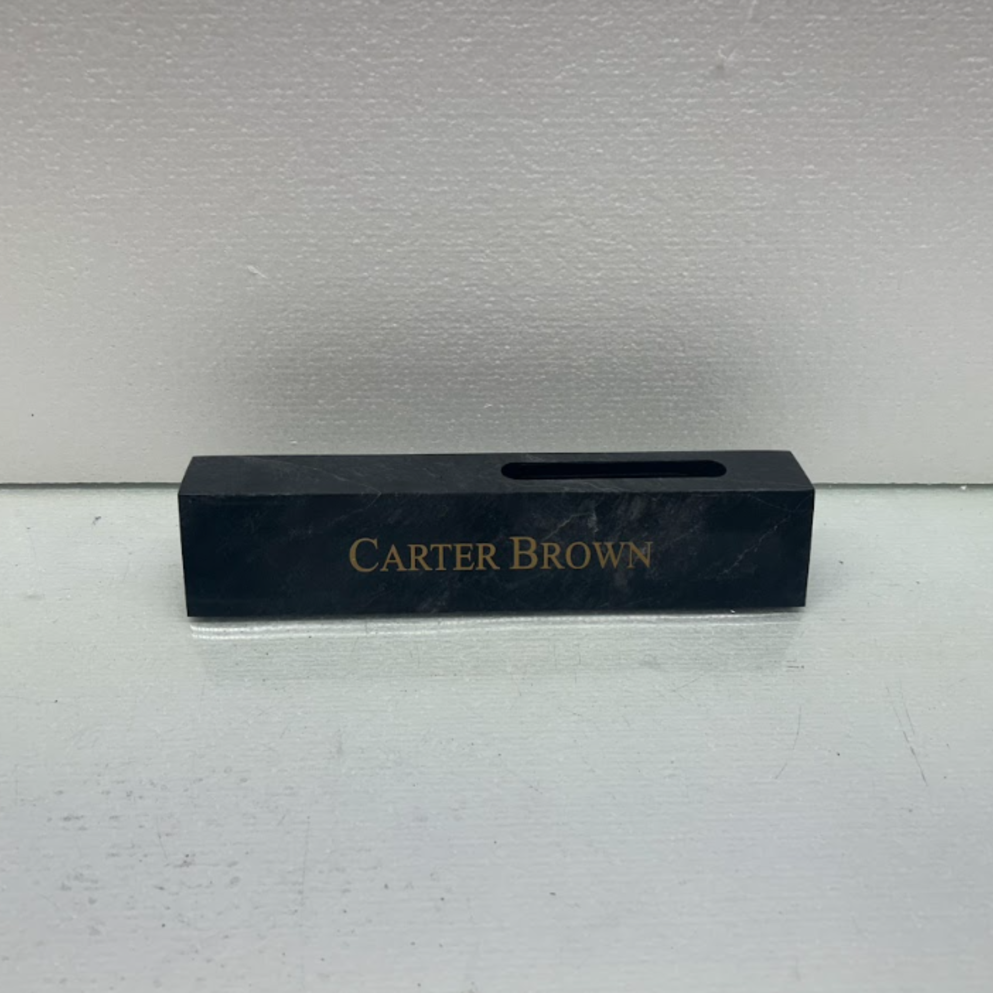 A black box with gold lettering that reads 'Carter Brown,' placed on a gray surface in front of a plain white wall.
