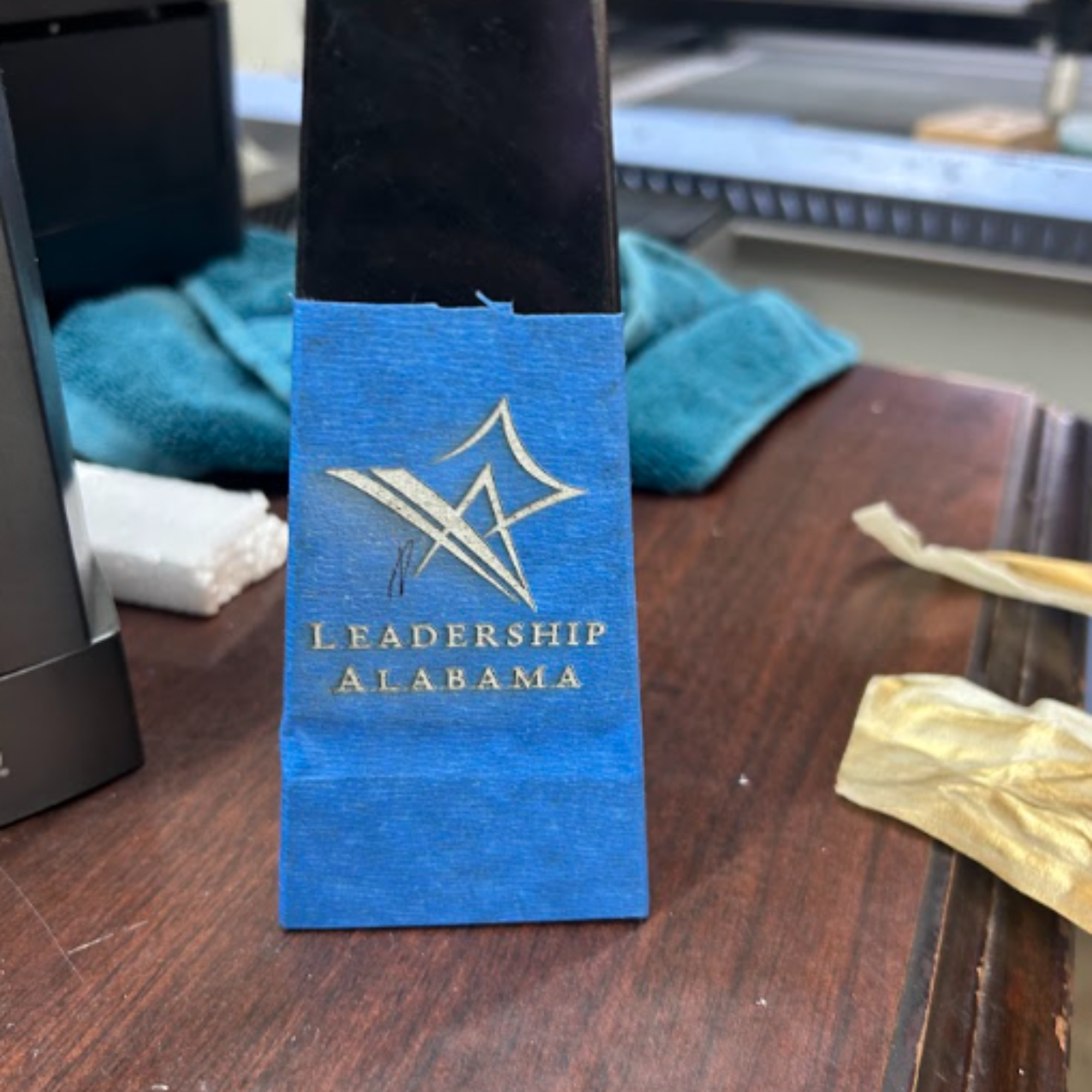 Blue lanyard with a gold mountain logo and the words "Leadership Alabama" on a wooden desk with some papers and electronic devices in the background.