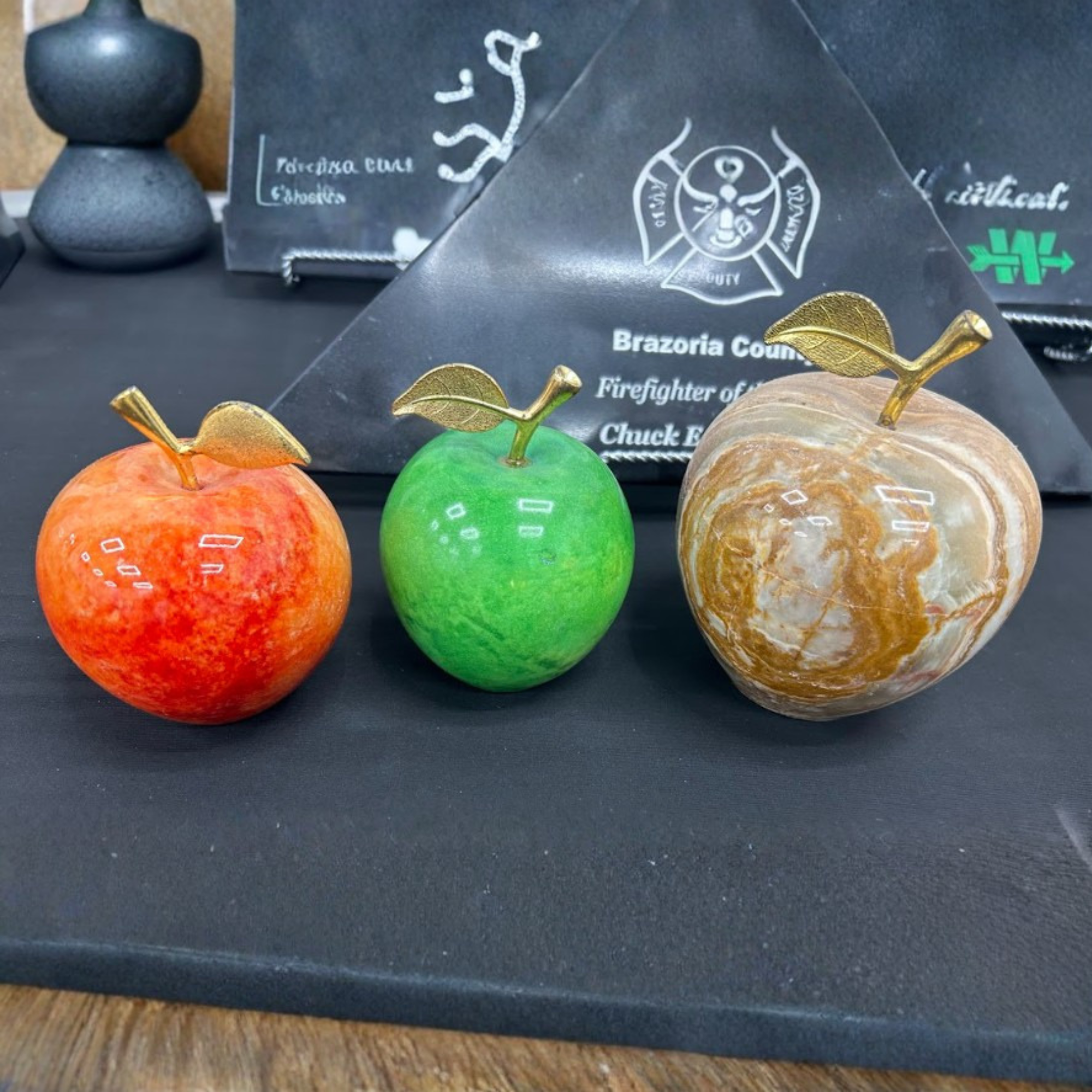 Close-up of decorative apples with gold leaves on top, placed on a dark surface, with parts of awards and plaques visible in the background.