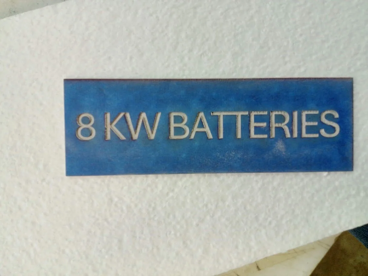 Blue sign with white 3D letters reading "8 KW BATTERIES" mounted on a textured white wall.
