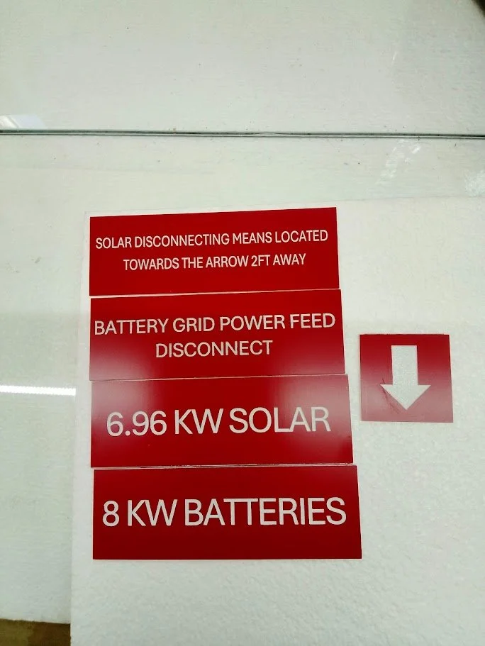 Red informational signs indicating solar and battery power details, including disconnect instructions, power feed, and capacity ratings.
