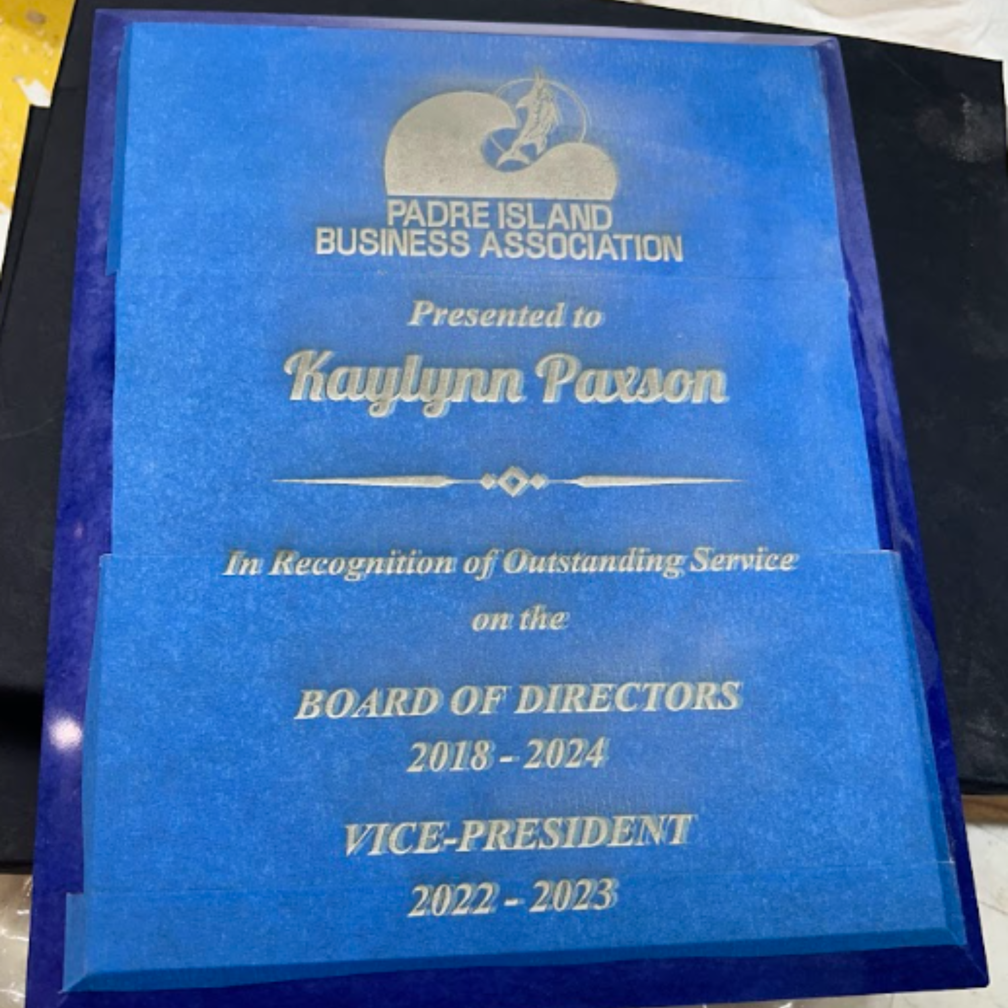 Close-up of a blue plaque awarded by the Padre Island Business Association recognizing Kaylinn Pavon for her outstanding service, with the years 2018 to 2024 visible.