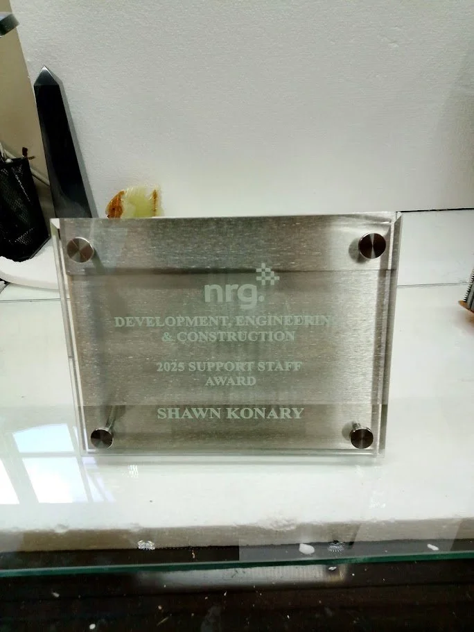 Metal plaque on a white surface, reading 'NRG Development, Engineering & Construction, 2025 Support Staff Award, Shawn Konary'.