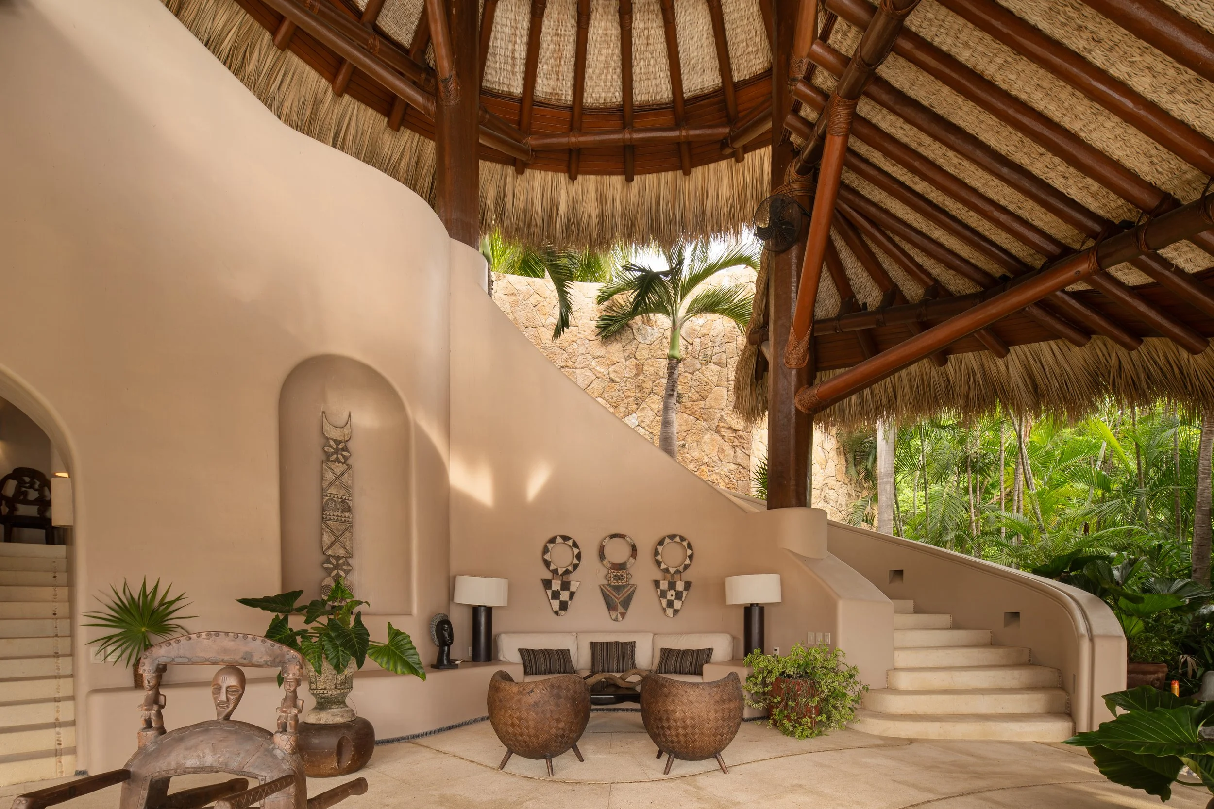 Interior of a tropical hotel lobby with a thatched roof, curved white wall, and tropical plants. Decorative tribal artwork hangs on the wall behind a seating area with woven chairs and striped pillows, flanked by white lamps and plants.