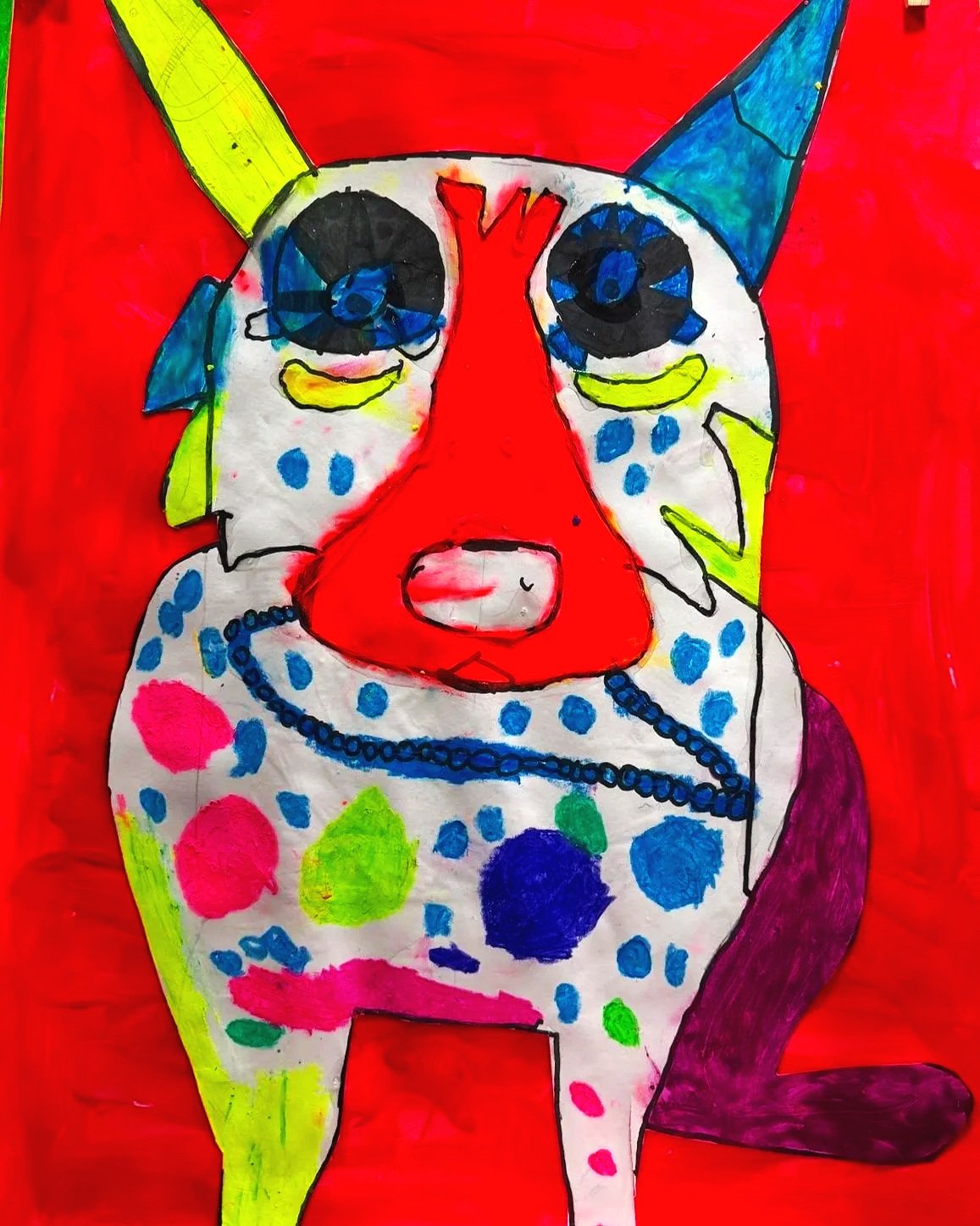 Colorful child’s drawing of a cat-dog creature with one yellow and one blue ear, a red nose, large black eyes, and a white body with multicolored spots, set against a red background.
