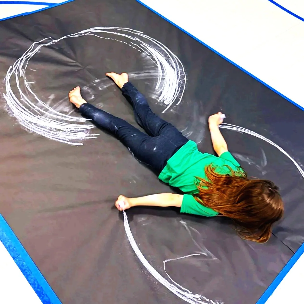 A young girl in a green shirt and black pants lying on a trampoline floor, creating chalk circles with her body and arms.