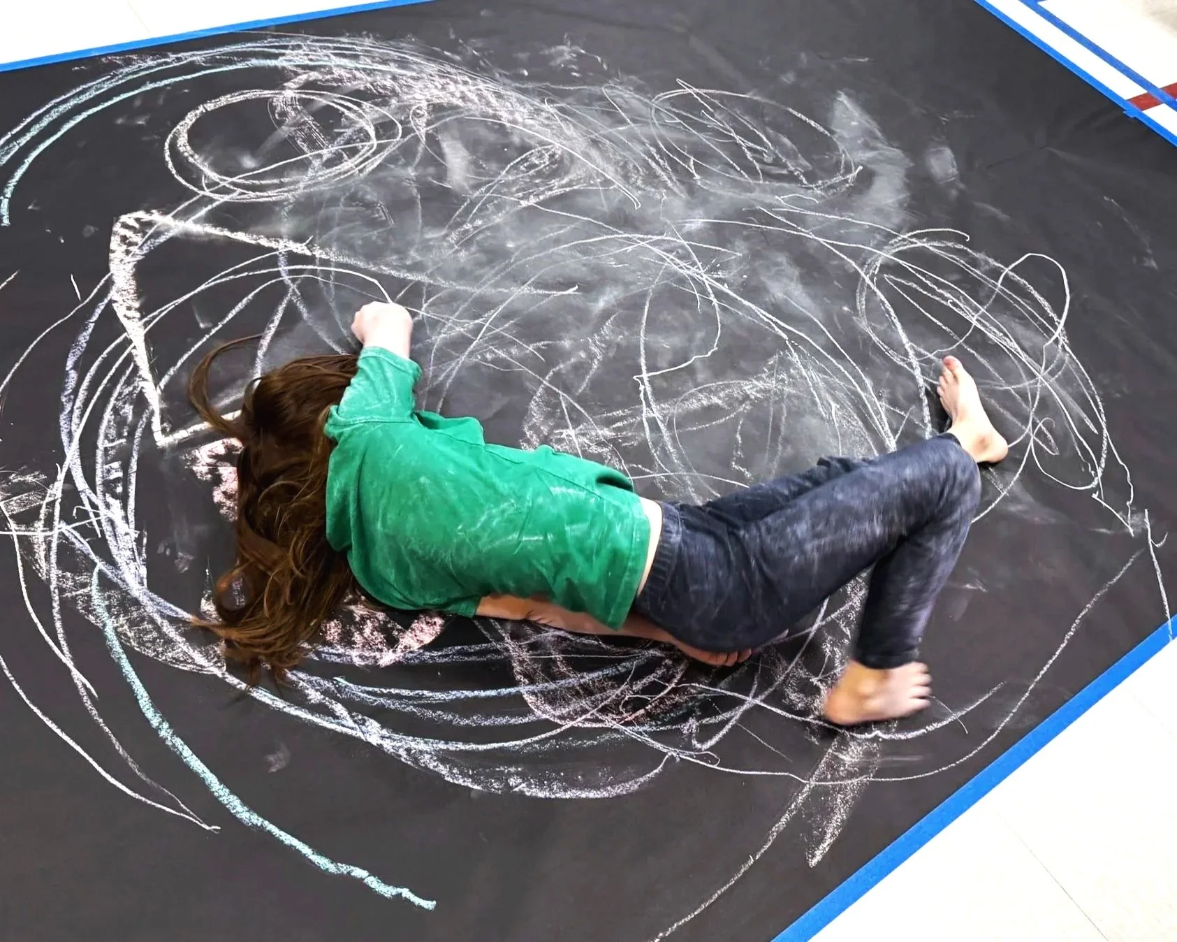 Child lying on large black mat drawing with white chalk, wearing a green shirt and dark pants, surrounded by colorful chalk marks.