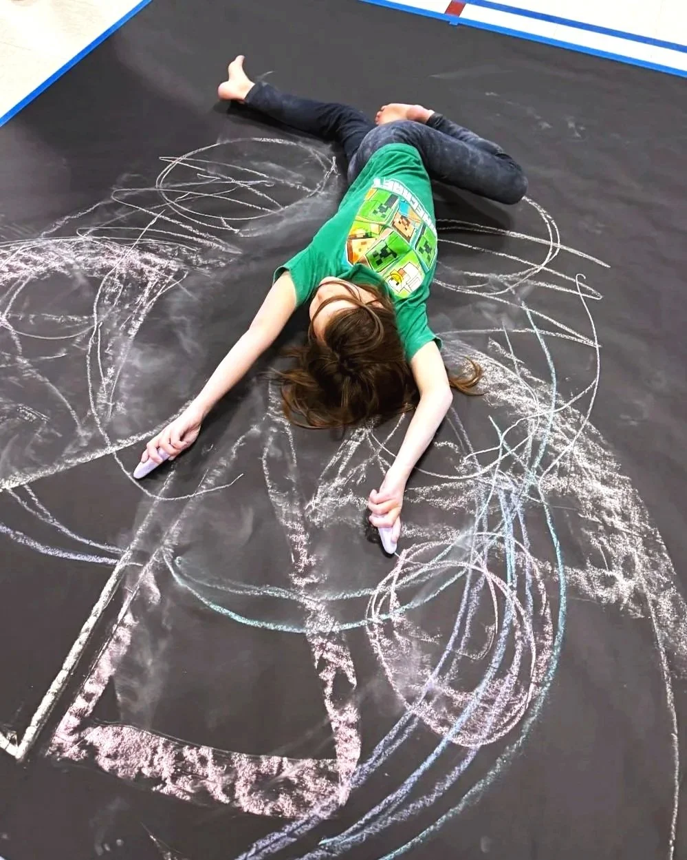 Child lying on a black chalkboard floor drawing with colored chalk, surrounded by white chalk scribbles.