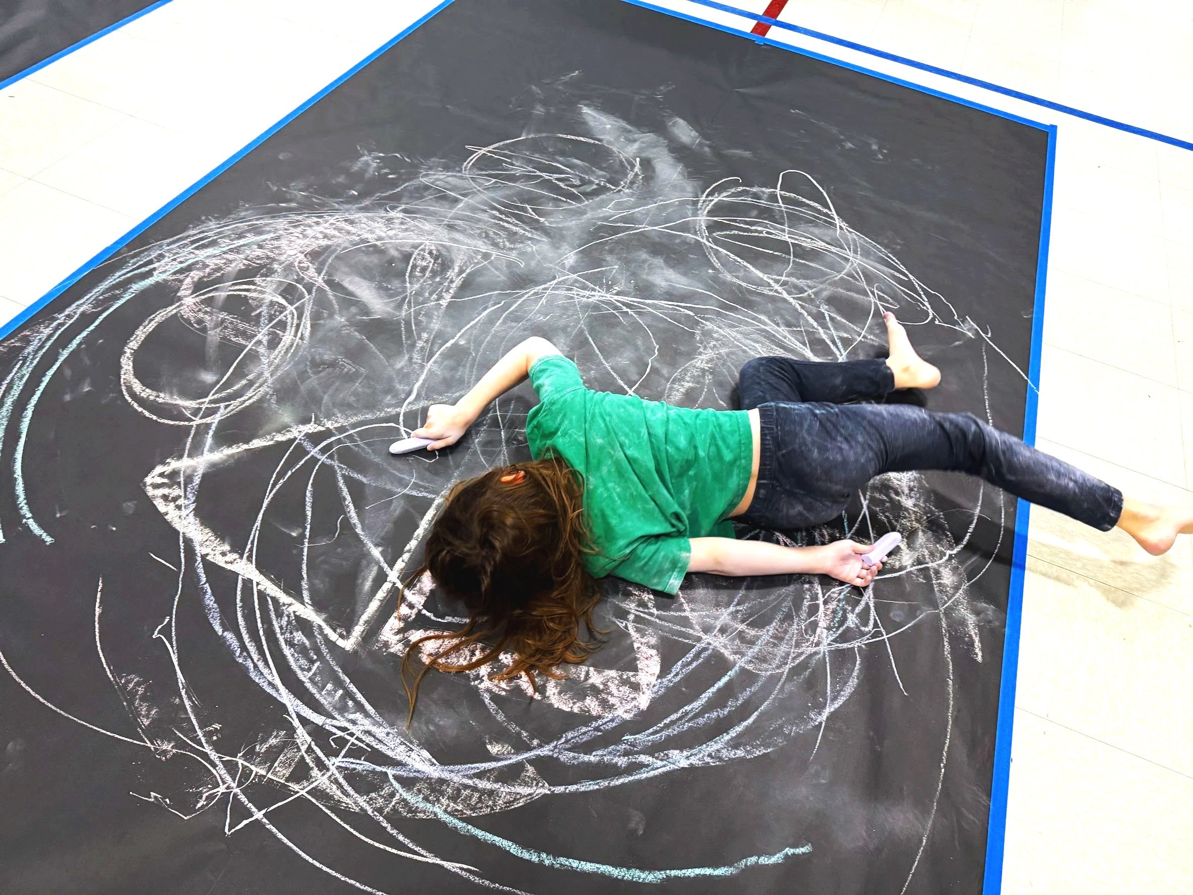 Child lying on a black chalkboard floor, drawing with white chalk in a spiral pattern, holding a cell phone in one hand.